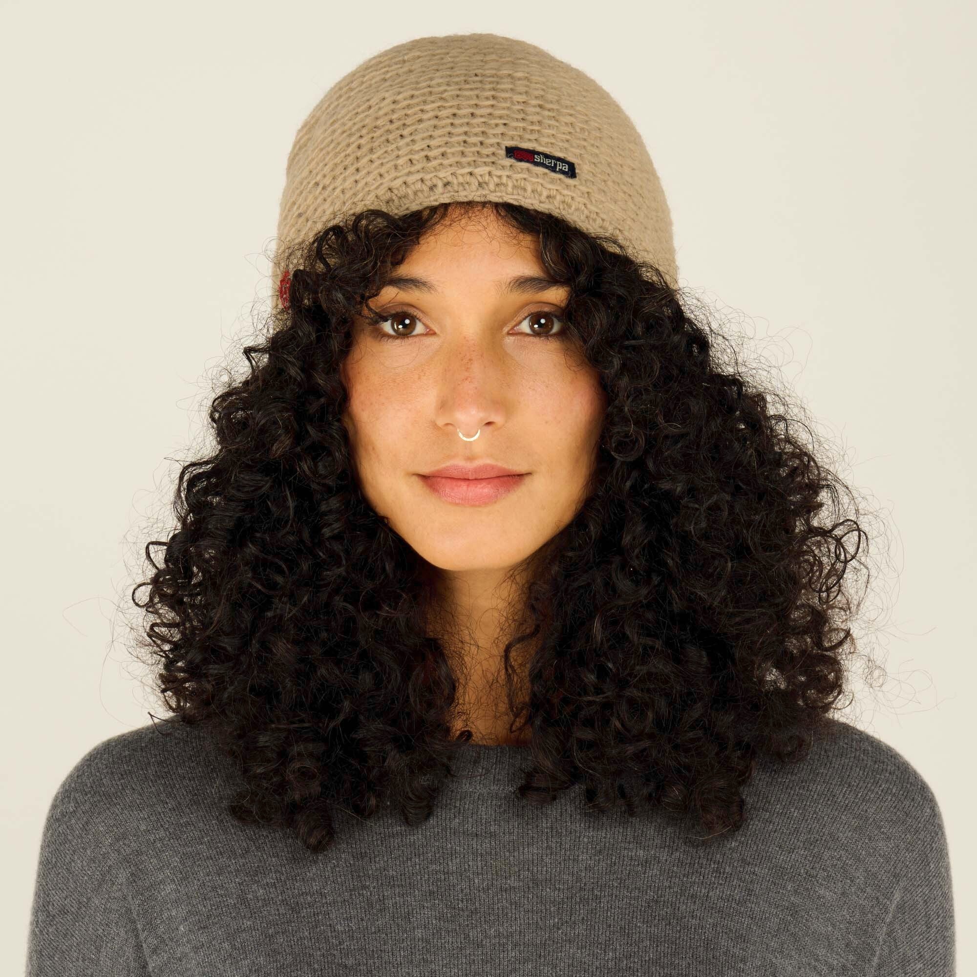 A front-facing view of a model wearing a Sherpa Adventure Gear Jumla Hat in Light Brown with a textured pattern. The beanie features a small Sherpa logo tag on the front. The model has curly hair and is dressed in a grey sweater against a neutral background, emphasising the beanie's details.