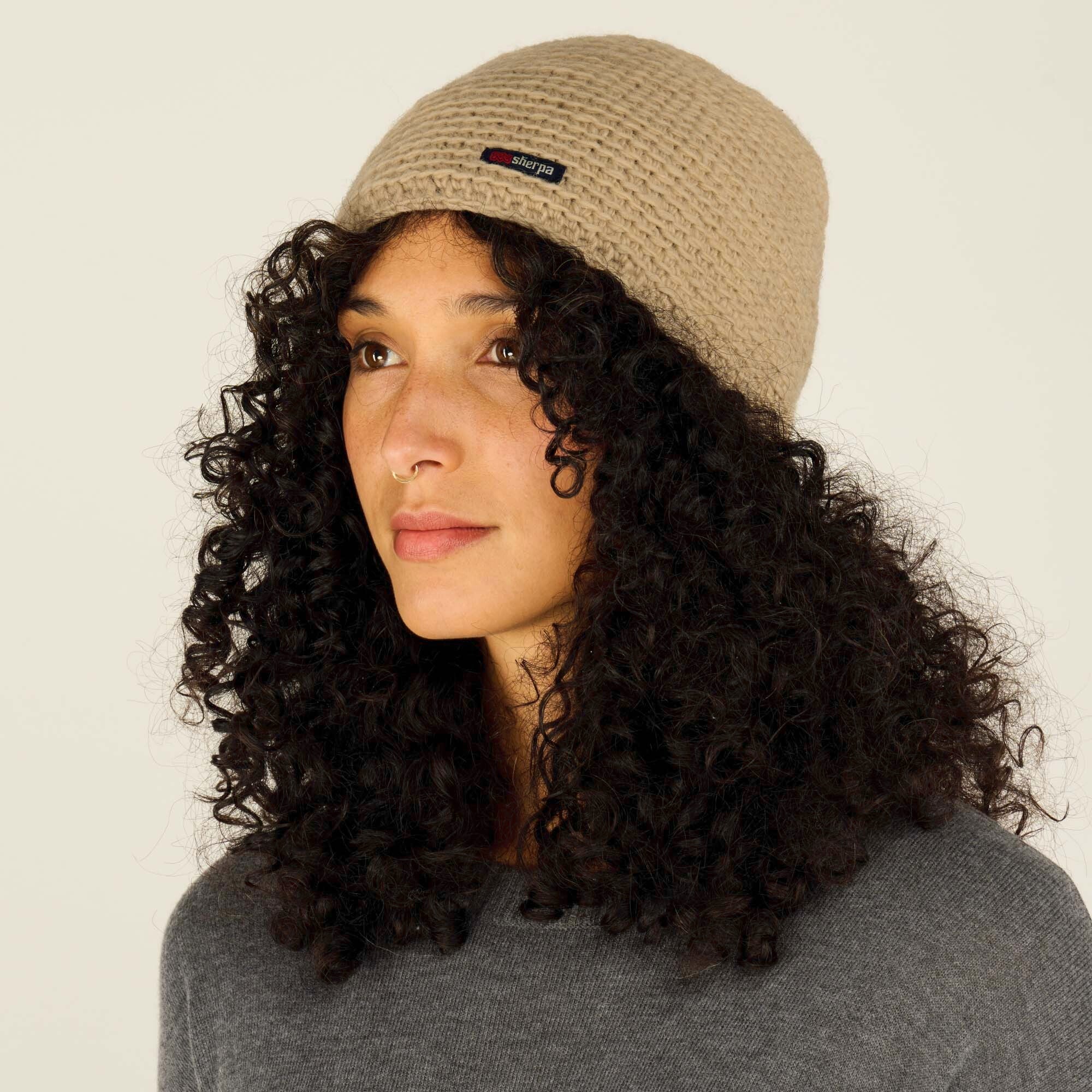 A side-profile view of a model showcasing a Sherpa Adventure Gear Jumla Hat in Light Brown with a textured design. The beanie prominently displays a small Sherpa logo tag. The model has voluminous curly hair and is dressed in a grey sweater, with the neutral background highlighting the beanie's texture and shape.