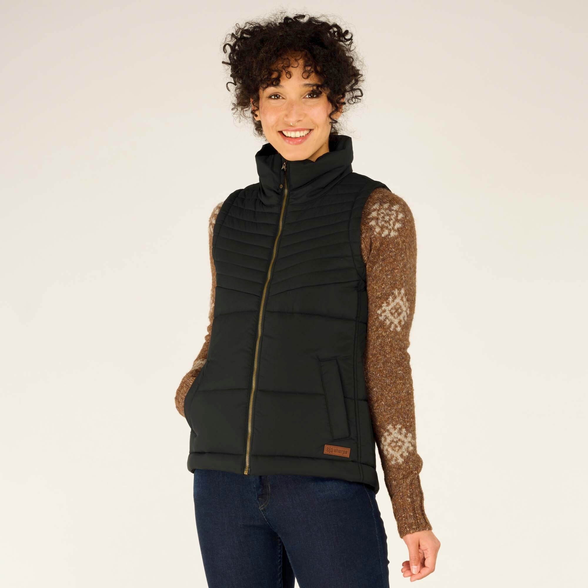 A smiling woman with curly hair models a Sherpa Adventure Gear Kabru Vest in Black over a brown patterned sweater. The vest has a high collar, a quilted design, and a full-length zipper. She places her hands in the vest's side pockets while looking slightly off-camera.