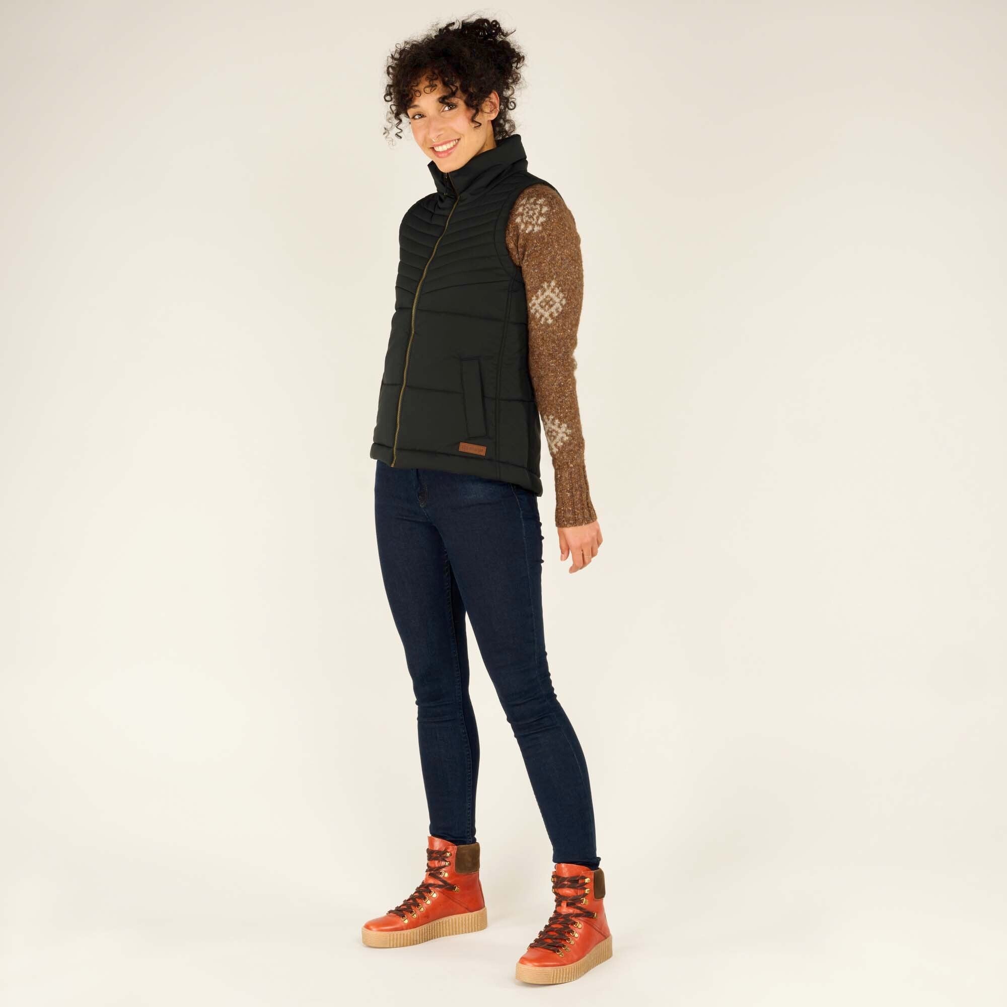 The model stands with one leg slightly bent, showcasing the Sherpa Adventure Gear Kabru Vest in Black styled with dark blue skinny jeans and tan high-top boots with red laces. The vest features a horizontal quilted pattern, high collar, and zippered front.