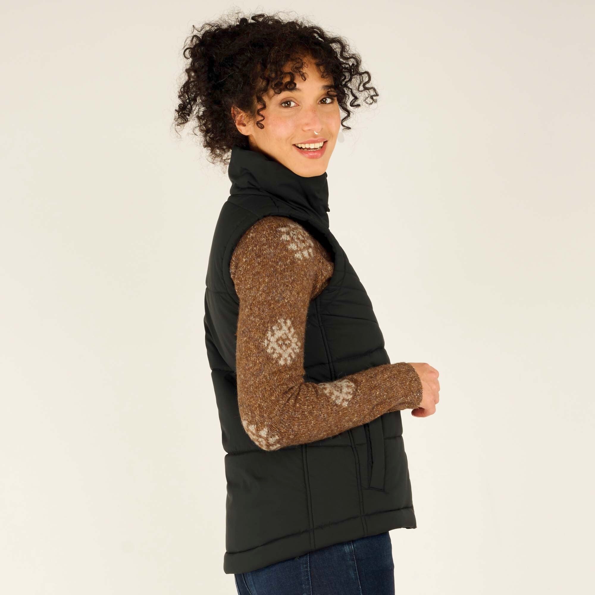 The model turns slightly to her left, revealing the Sherpa Adventure Gear Kabru Vest in Black’s fitted silhouette. The vest has a smooth quilted finish and is layered over a brown knit sweater with white geometric patterns on the sleeves.