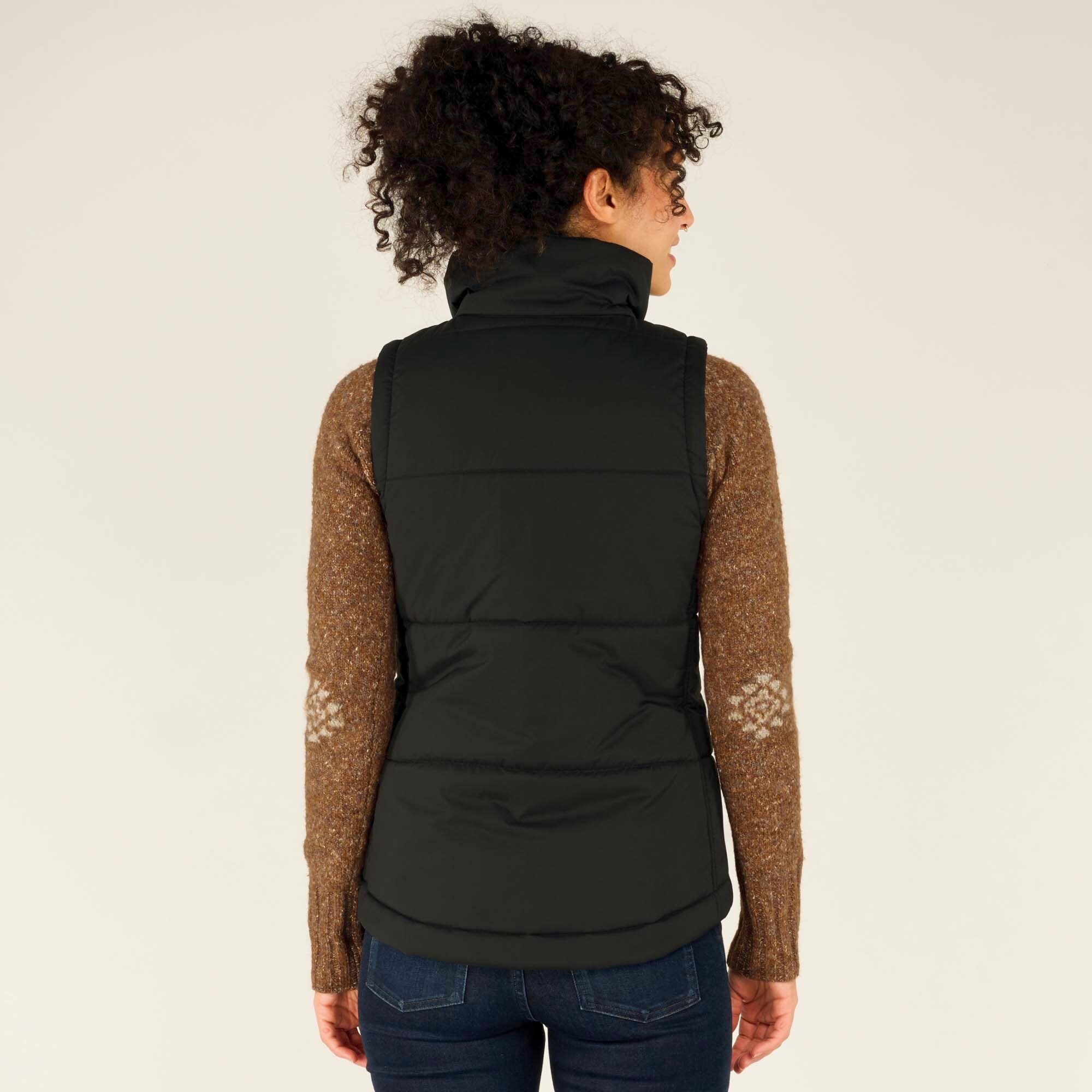 A rear view of the model showcases the back of the Sherpa Adventure Gear Kabru Vest in Black, which has a structured fit and horizontal quilted design. The high collar and slightly tapered waist are visible, along with the brown patterned sweater peeking from underneath.