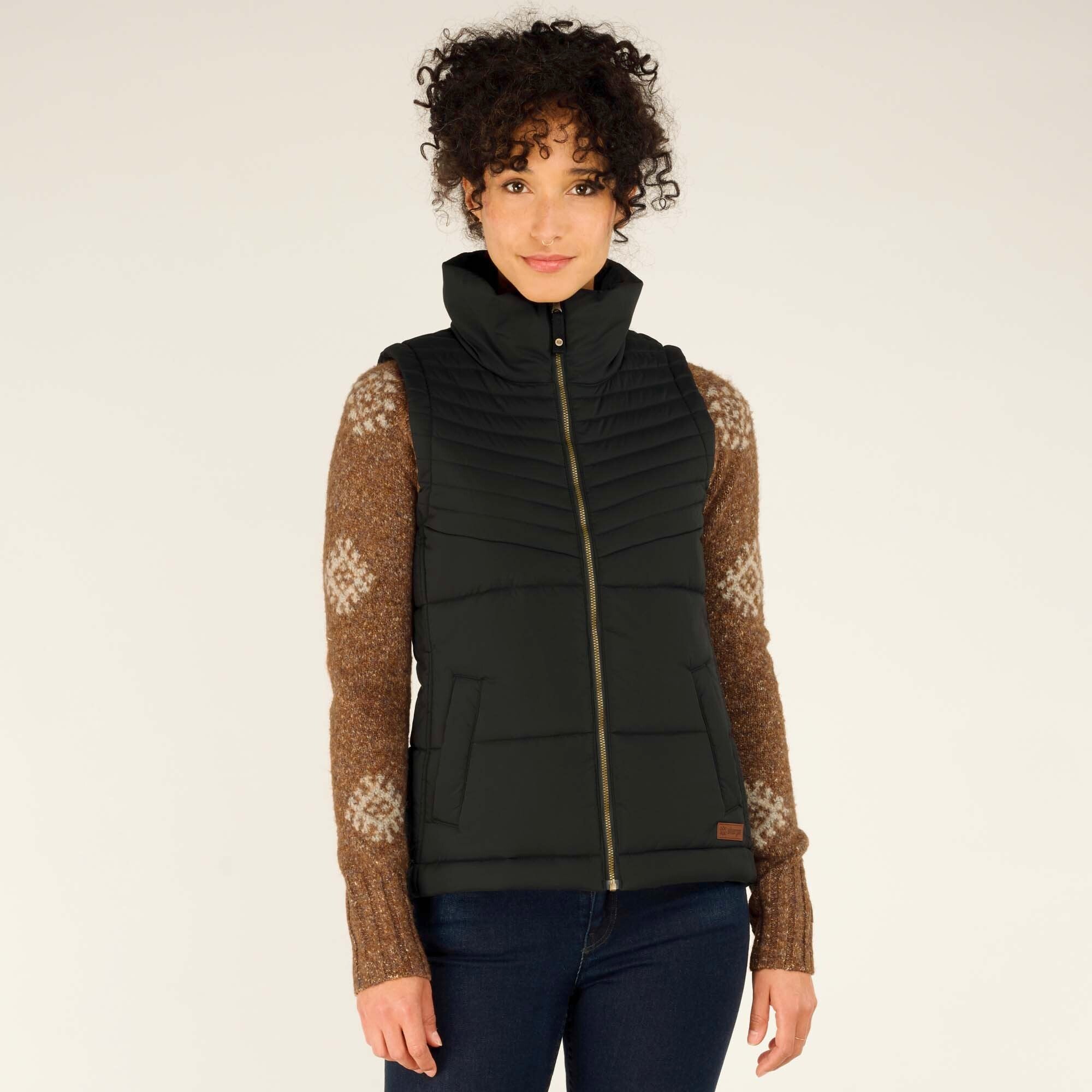 The model stands facing the camera, looking directly at it with a subtle smile. The full-length zipper and structured design of the Sherpa Adventure Gear Kabru Vest in Black are clearly visible. The brown sweater’s white pattern on the arms contrasts with the sleek black vest.