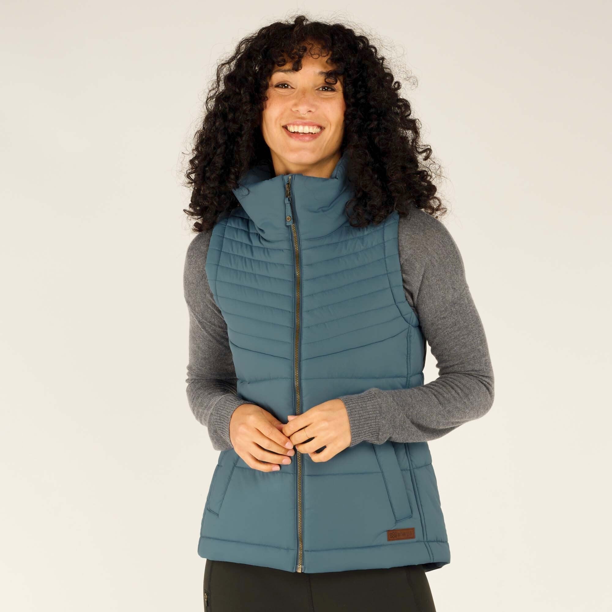 A woman wearing a Sherpa Adventure Gear Kabru Vest in Blue over a gray long-sleeve top. The vest features a high collar, zipper closure, and subtle branding near the hem. She is smiling and has her hands clasped in front.