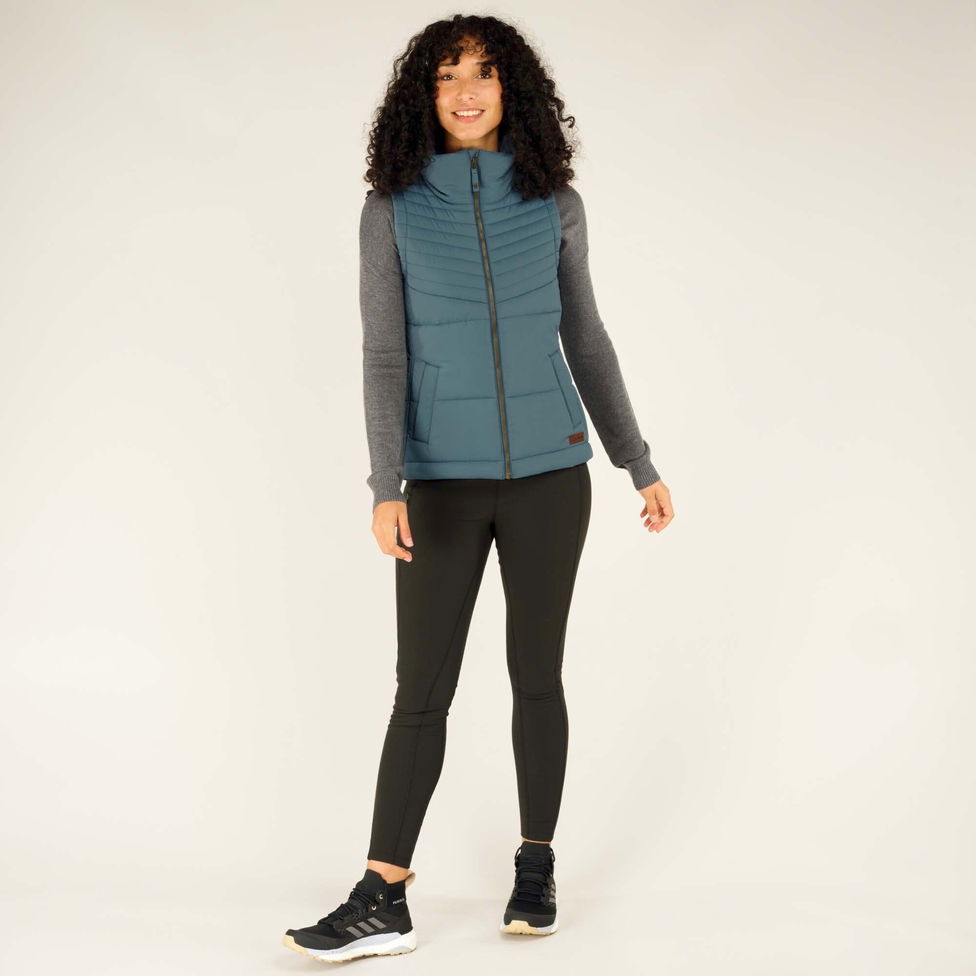 Full-body image of the same woman dressed in a Sherpa Adventure Gear Kabru Vest in Blue paired with dark leggings and black trainers. She stands confidently, smiling, in a relaxed pose.