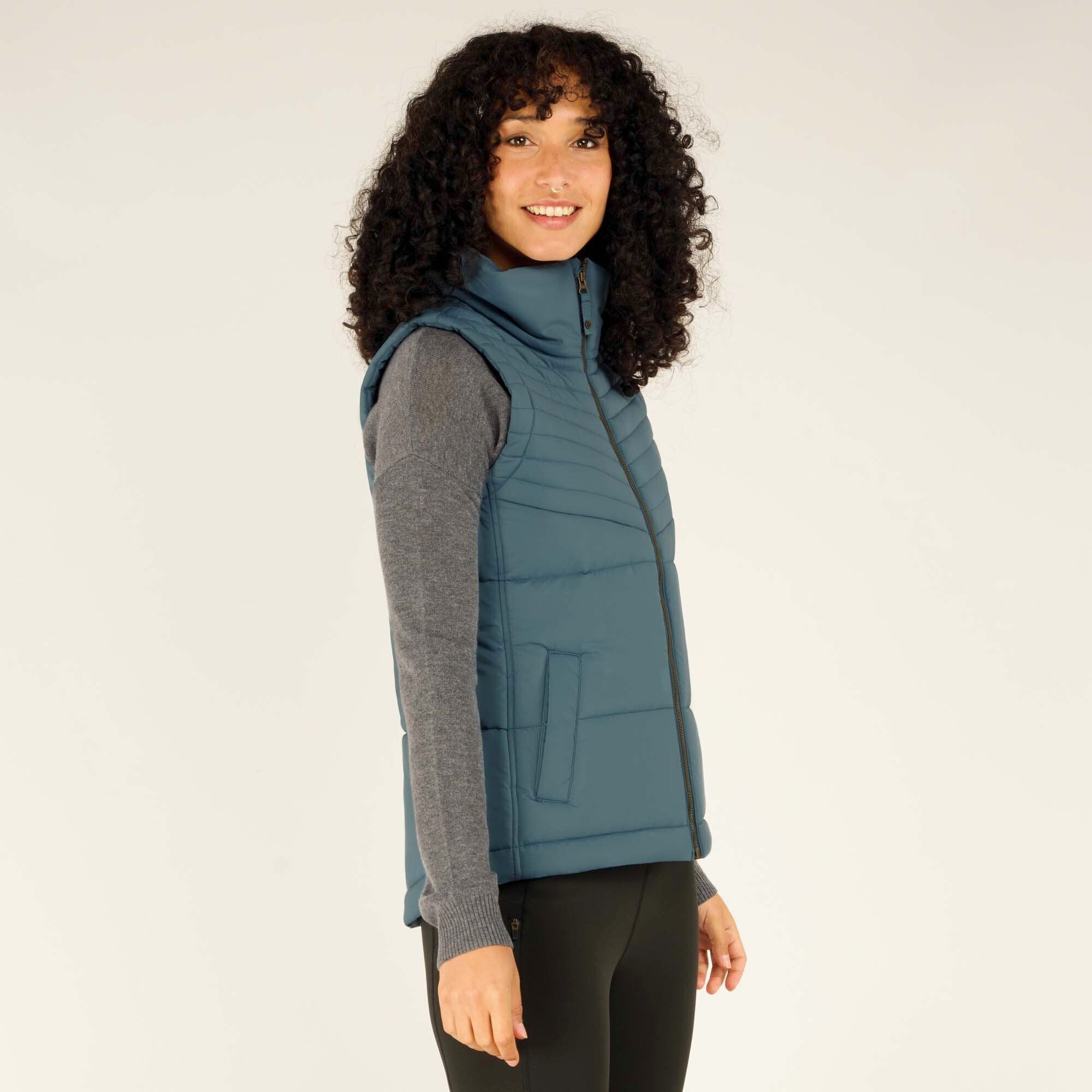 Side profile of the woman wearing the Sherpa Adventure Gear Kabru Vest in Blue. The quilting detail and fitted silhouette of the vest are highlighted, while she smiles warmly.