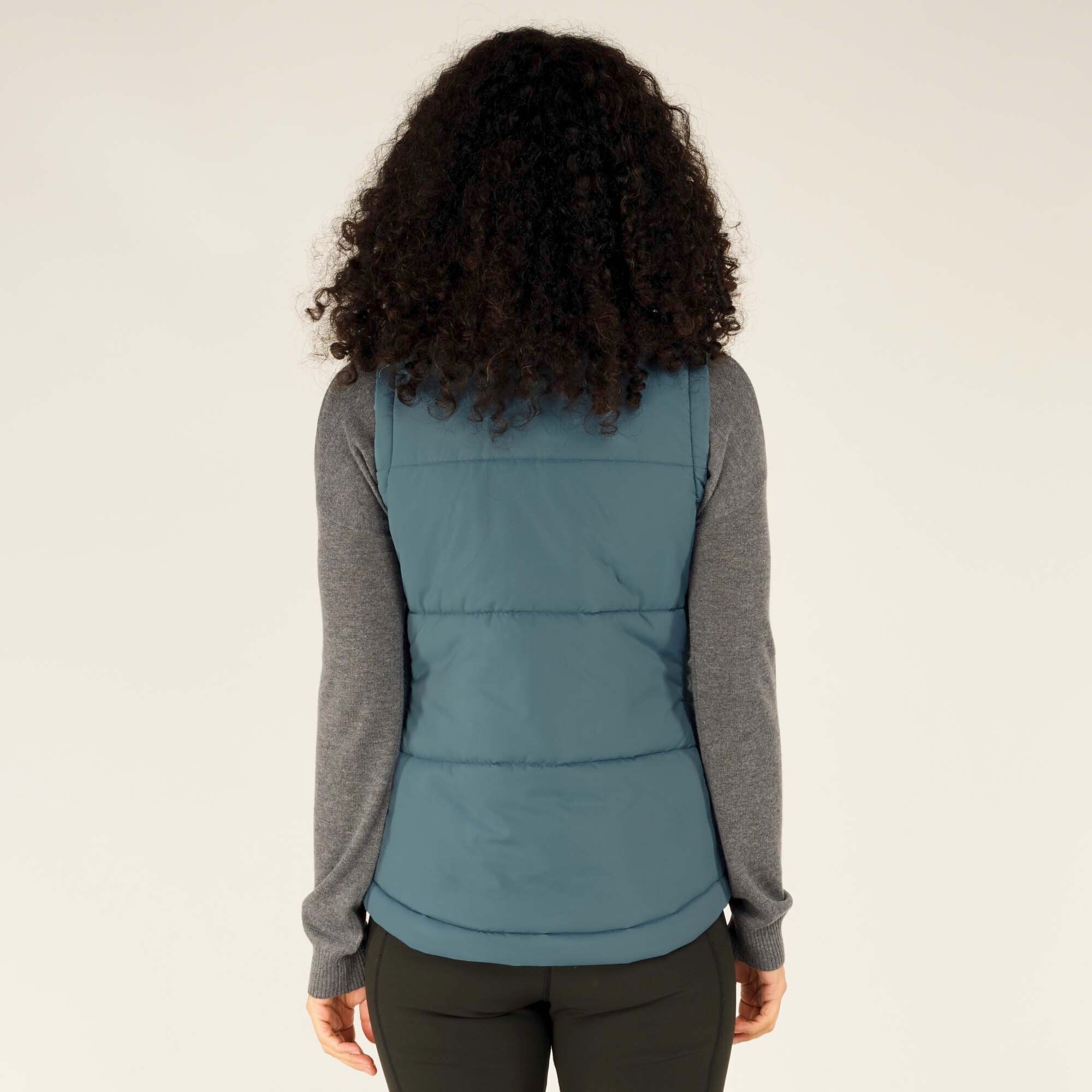 Rear view of the Sherpa Adventure Gear Kabru Vest in Blue, showing the clean design and horizontal stitching. The woman has her back to the camera with her curly hair framing the vest.