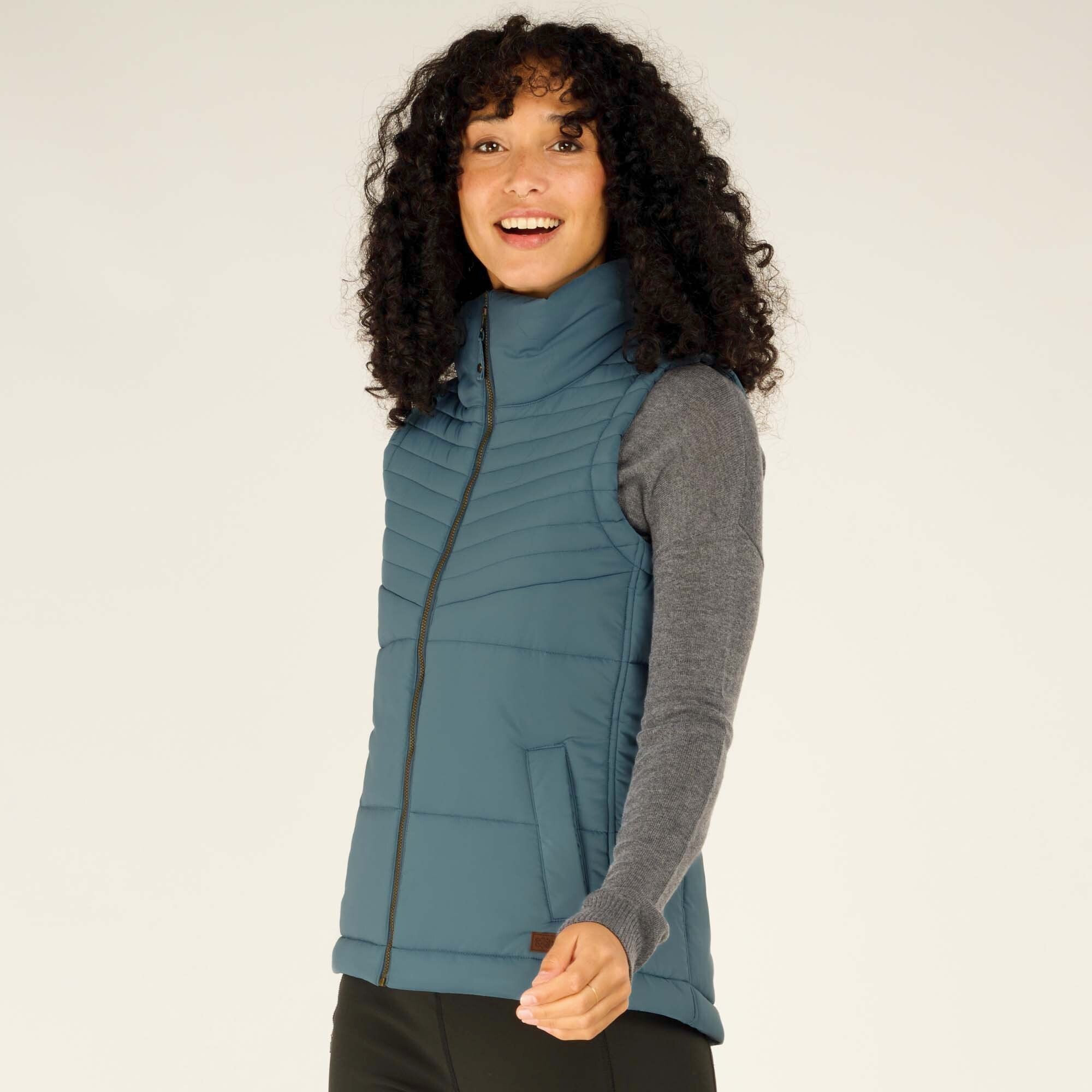 The woman turns slightly while smiling, showcasing the Sherpa Adventure Gear Kabru Vest in Blue from an angled view. The high collar and tailored fit are prominently displayed.