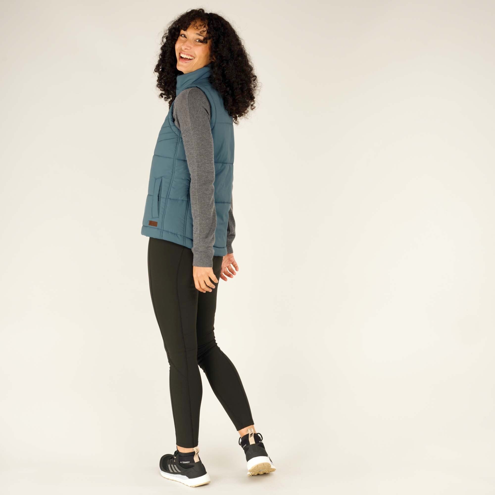 The woman in a playful stance, turning back slightly while smiling. She wears the Sherpa Adventure Gear Kabru Vest in Blue with dark leggings and black trainers, emphasising the versatility of the outfit for outdoor activities.