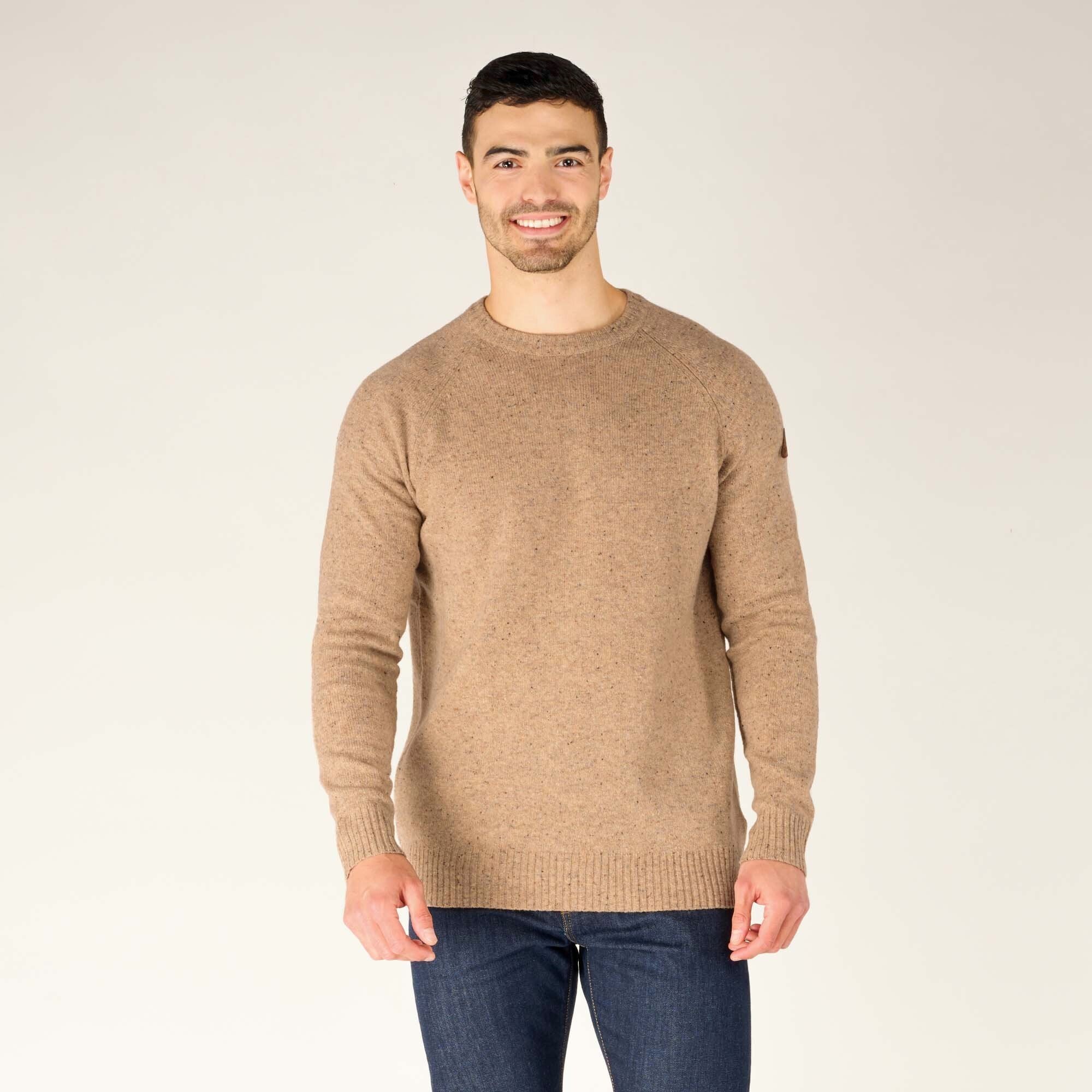 A male model with short dark hair and a beard is smiling at the camera while wearing a Sherpa Adventure Gear Kangtega Crew Sweater in Beige with a subtle speckled pattern. The sweater has ribbed cuffs, hem, and collar. He is also wearing dark blue jeans and stands in front of a neutral beige background.