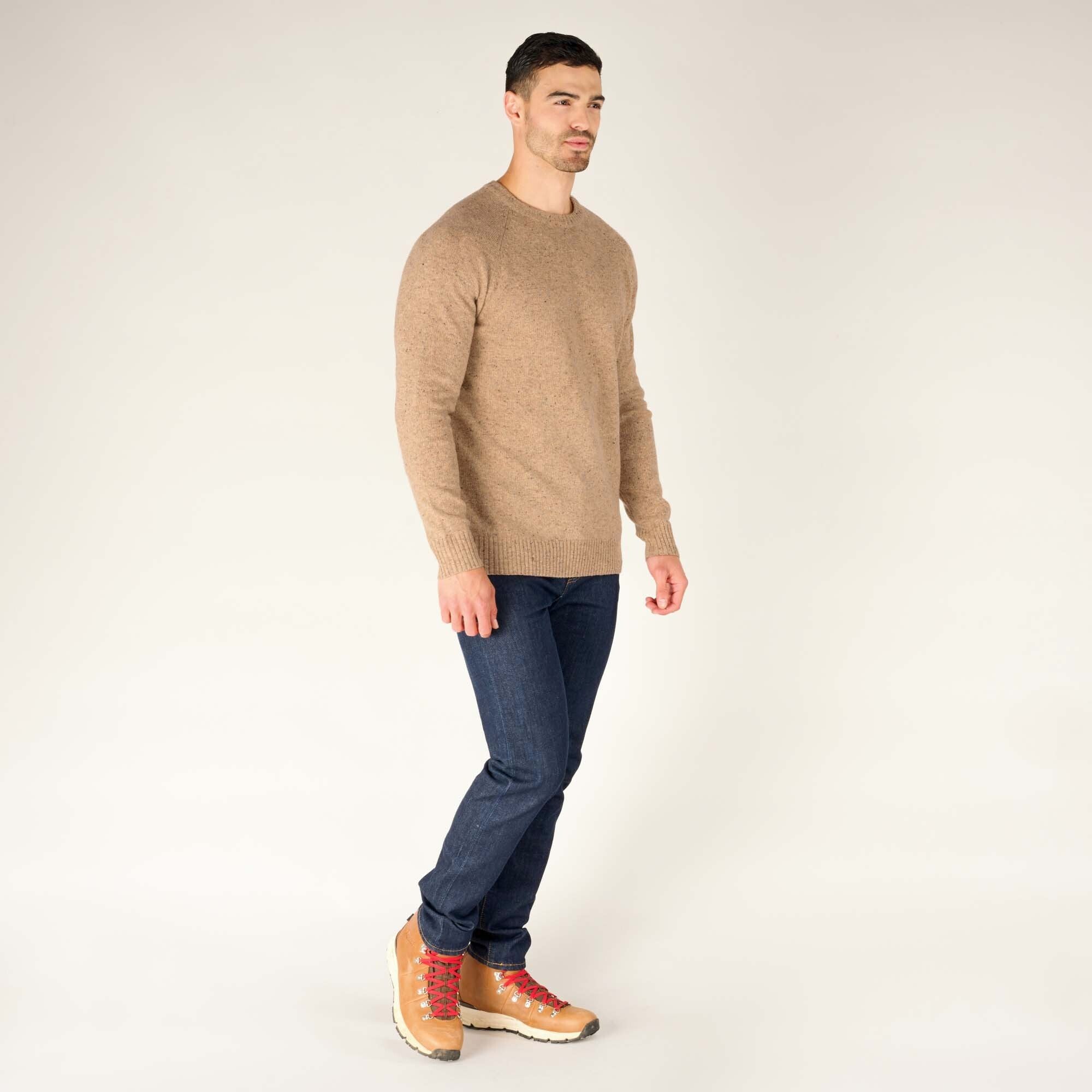 The same male model is shown in a full-body shot, walking slightly to the side while wearing the Sherpa Adventure Gear Kangtega Crew Sweater in Beige and dark blue jeans. He pairs the outfit with tan hiking boots that have red laces.