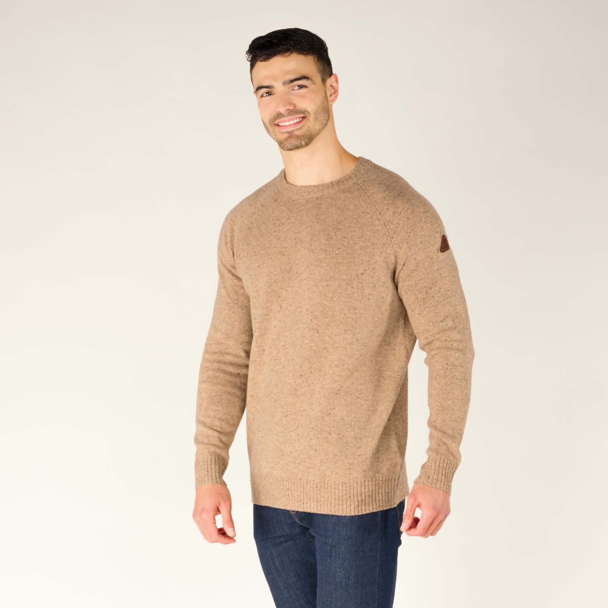 A three-quarter profile view of the male model wearing the Sherpa Adventure Gear Kangtega Crew Sweater in Beige, turned slightly to his left. He smiles while looking at the camera, showing the textured knit details of the sweater and a small leather brand patch on the upper left sleeve.