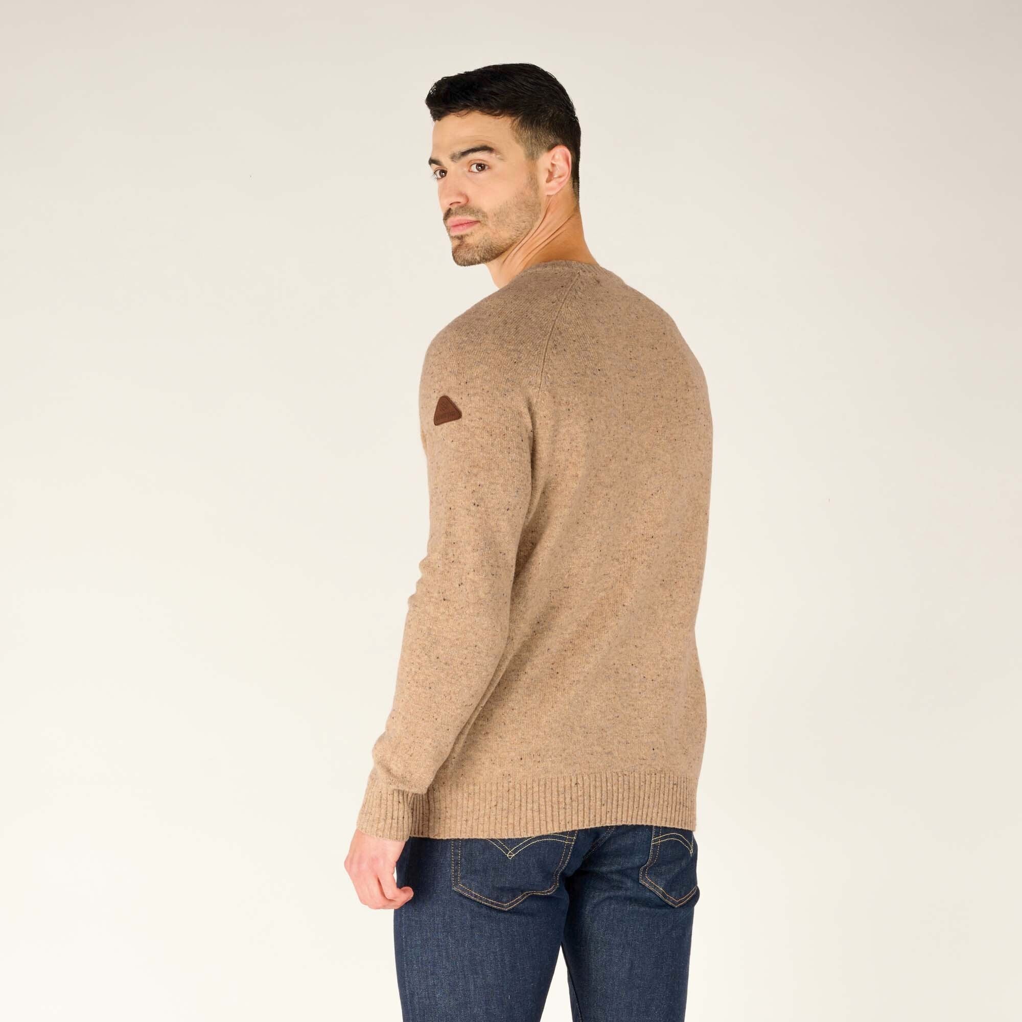 A side-back angle of the male model, looking over his left shoulder while slightly turning his body. This angle highlights the fit of the Sherpa Adventure Gear Kangtega Crew Sweater in Beige along the arms and torso, as well as the brand's leather patch on the sleeve. He maintains a neutral expression with a hint of a smirk.