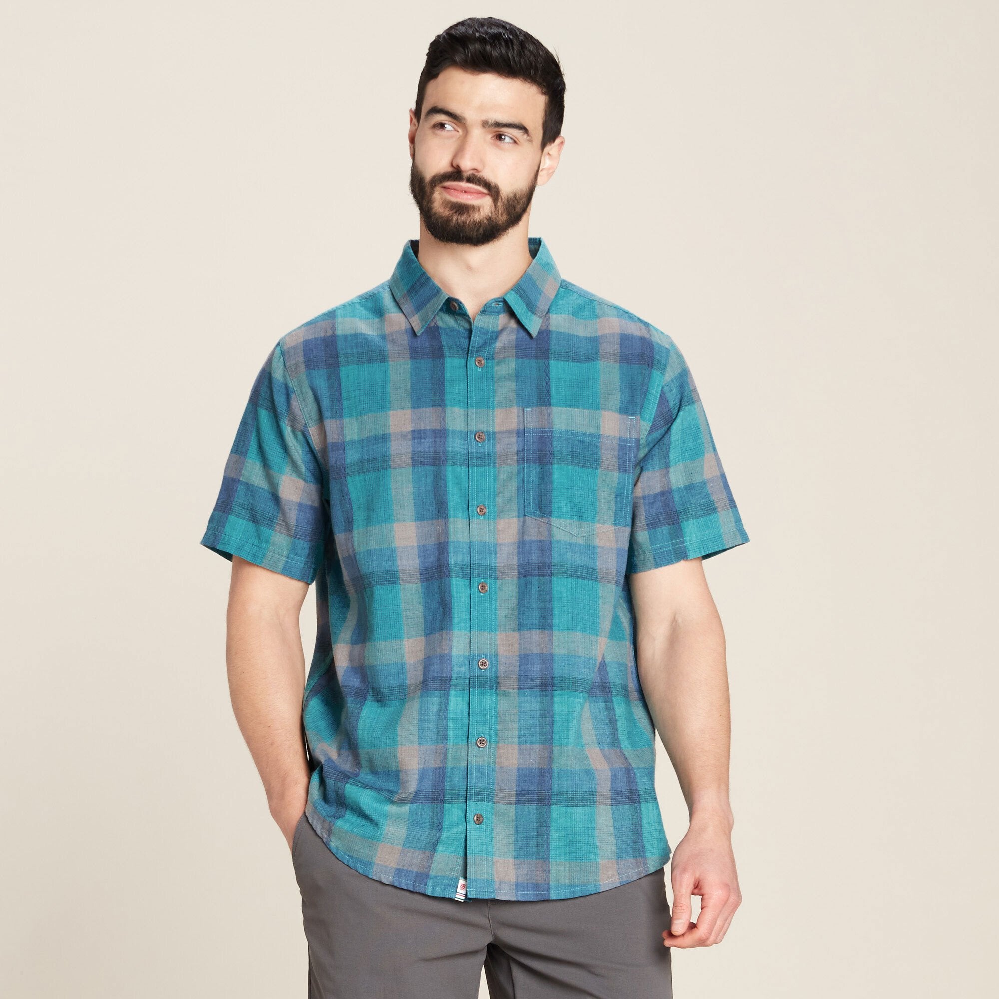 A man wearing a Sherpa Adventure Gear Khamsu Short Sleeve Shirt in Blue stands confidently, looking to the side. The shirt features a classic button-down design with a single chest pocket. The man pairs the shirt with grey pants, creating a casual yet polished look.