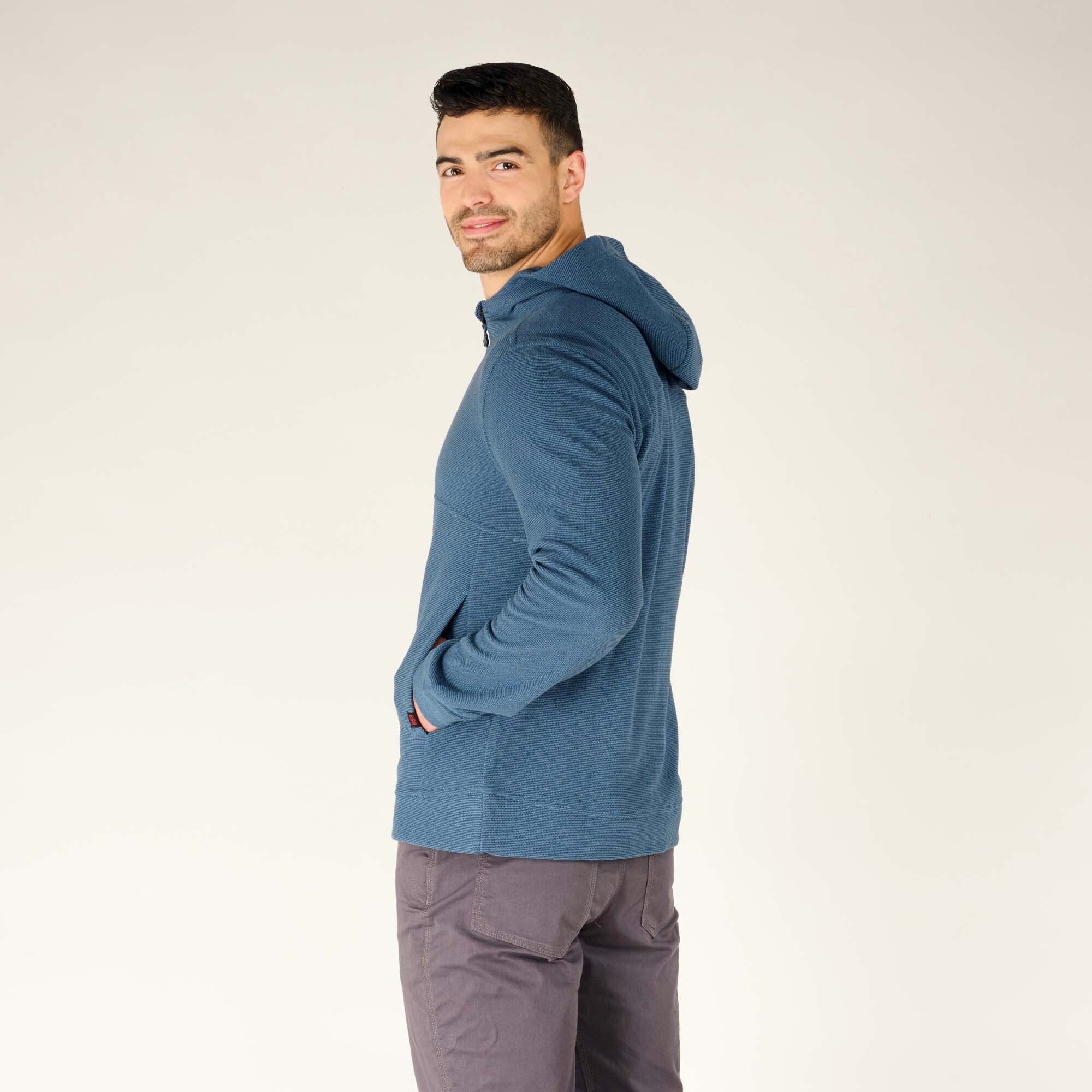 The man standing sideways and looking over his shoulder, wearing the Sherpa Adventure Gear Kula Full Zip Hoodie in Blue. His casual stance highlights the hoodie’s comfortable fit and the utility of the side pockets.