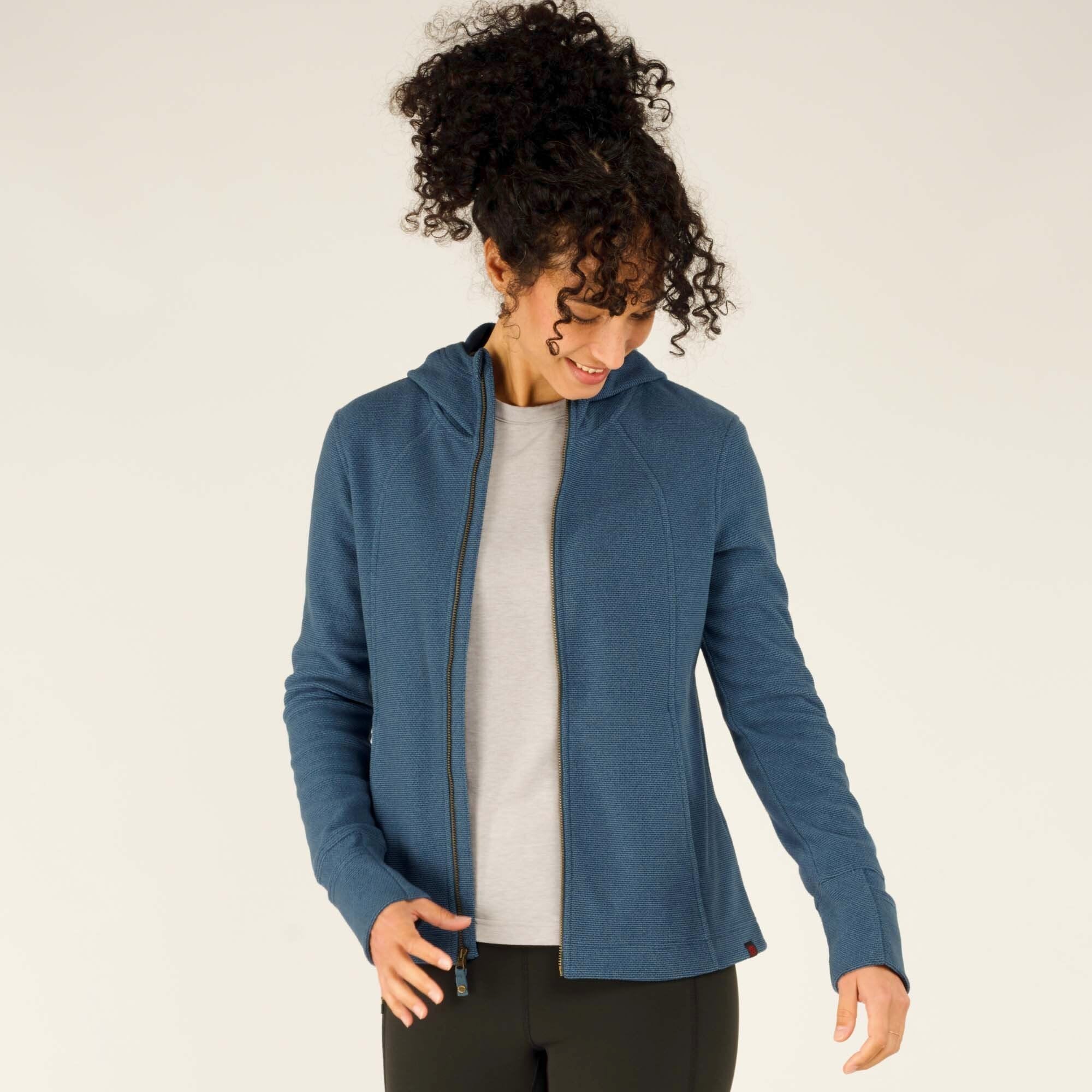 A woman with curly hair and a smile is wearing a Sherpa Adventure Gear Kula Full Zip Hoodie in Blue with a hood, a gray t-shirt underneath, and black leggings. She is unzipping the hoodie slightly while looking down and smiling. The background is a plain, light-colored studio backdrop.