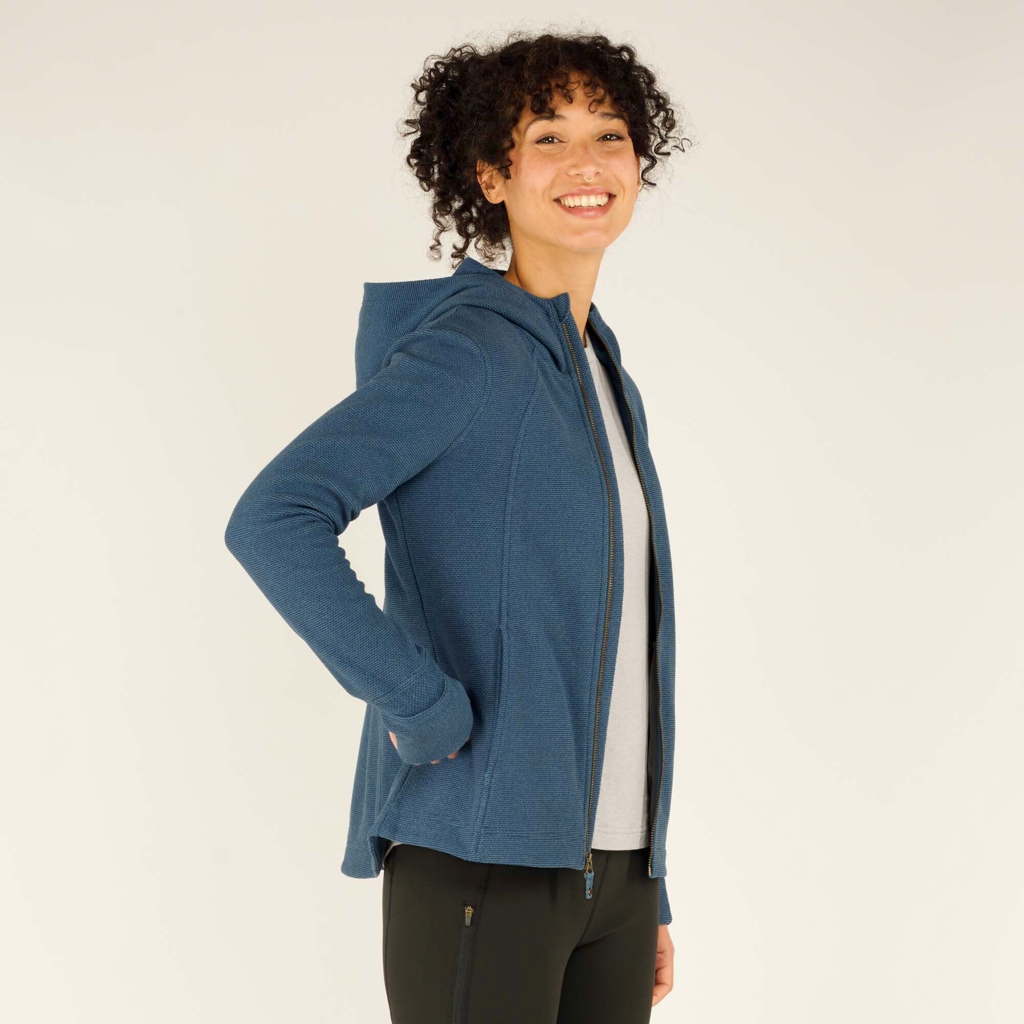 A side-angle image of the woman smiling with her hands in the Sherpa Adventure Gear Kula Full Zip Hoodie in Blue's pockets. The zippered hoodie is partially unzipped, revealing the gray t-shirt underneath. The hood is down, and the sleeves extend to her wrists with thumbholes.