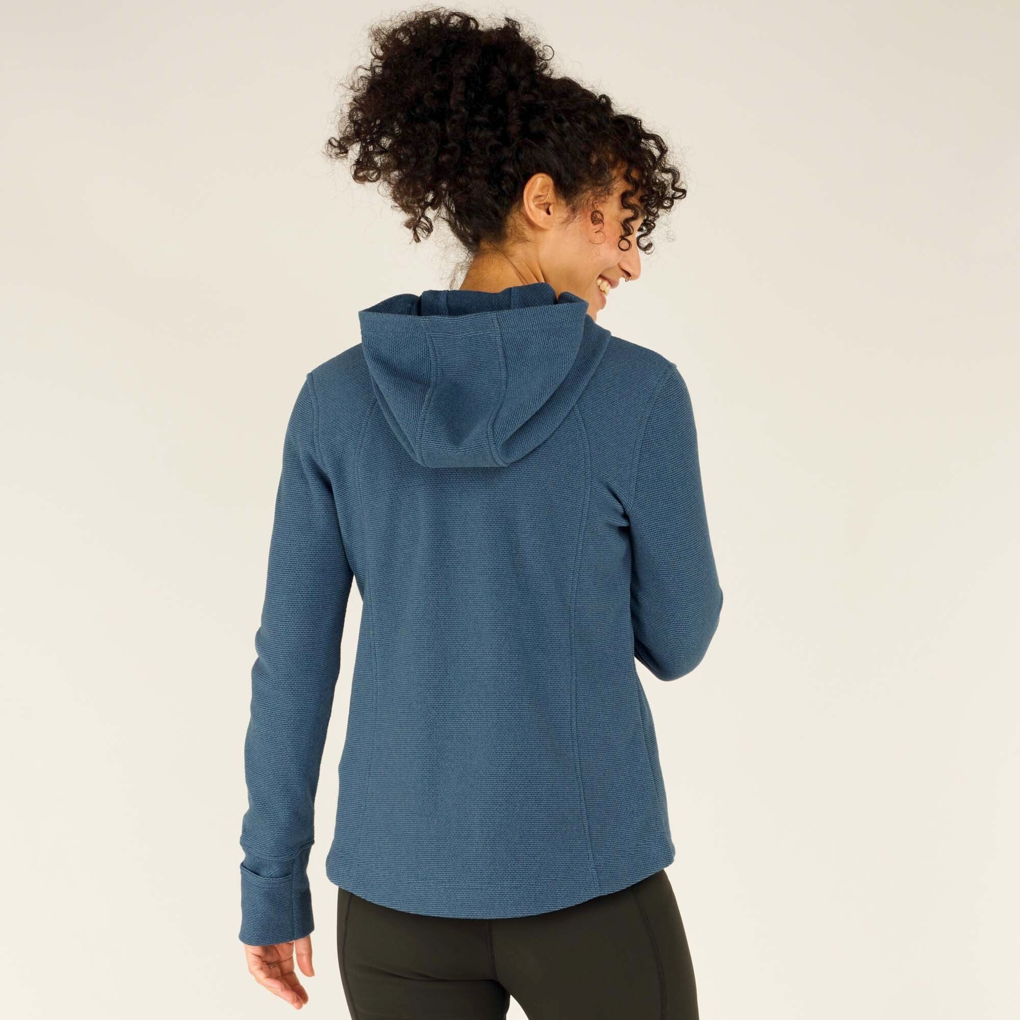 A back view of the woman wearing the Sherpa Adventure Gear Kula Full Zip Hoodie in Blue, with the hood up. She turns her head slightly to the side, smiling. The hood is structured, and the sleeves fit snugly along her arms.