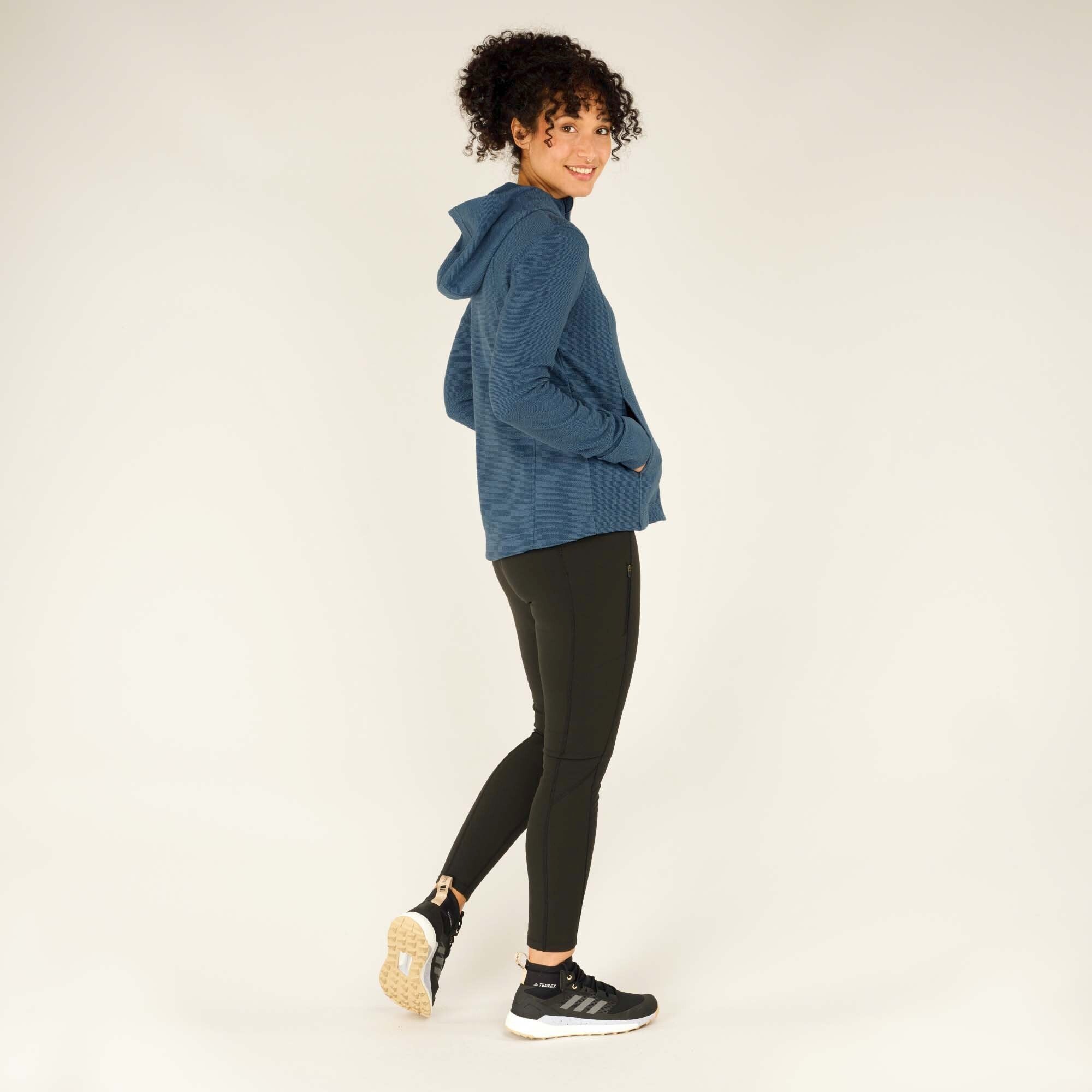 The woman is captured walking forward slightly, turning her head back towards the camera while smiling. The Sherpa Adventure Gear Kula Full Zip Hoodie in Blue is fully zipped, and the black leggings and sneakers give a sporty, active appearance.