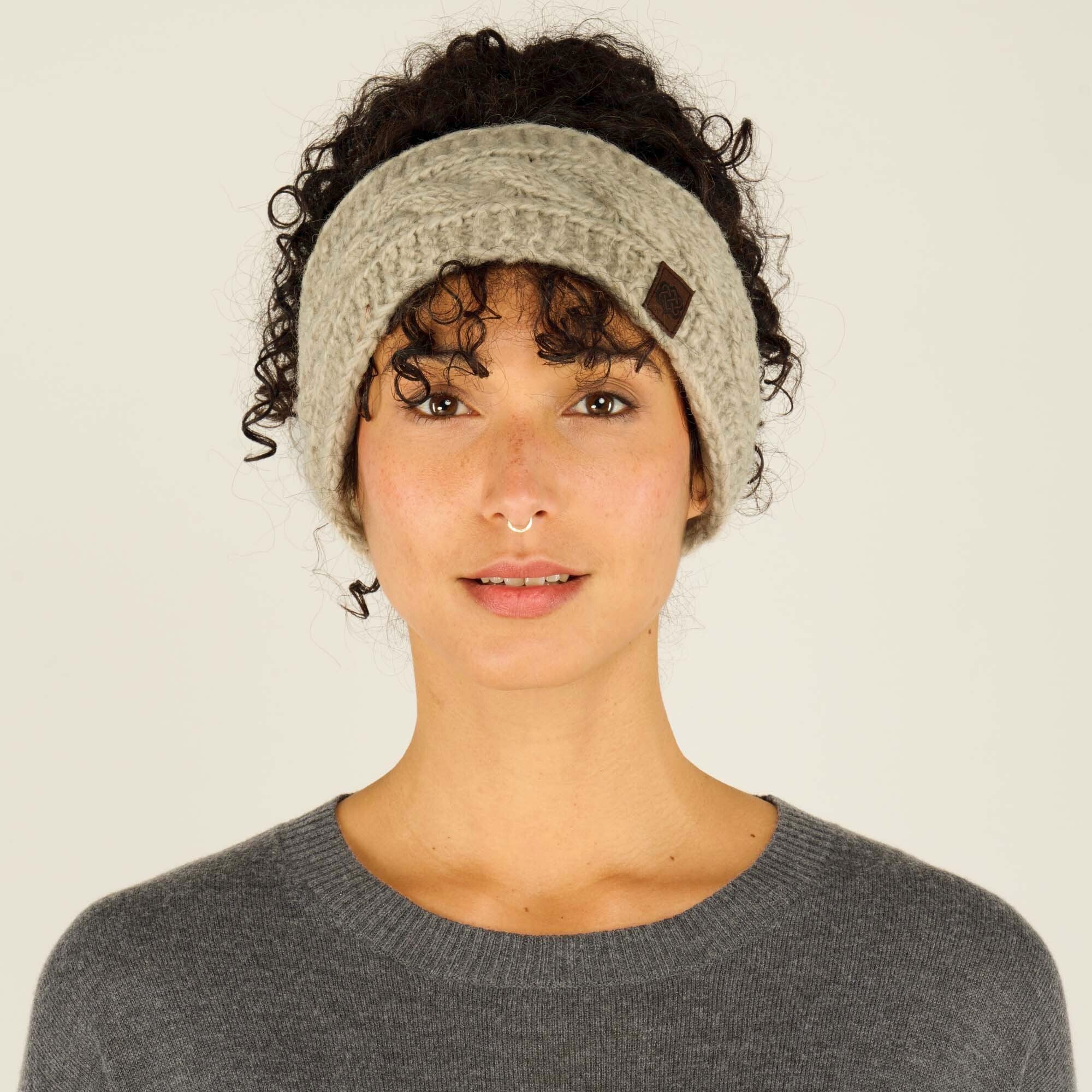 A woman with curly dark hair and a warm smile wears a thick, knitted Sherpa Adventure Gear Kunchen Headband in Light Brown with a cable-knit pattern. The headband has a small, square, brown leather patch with a logo stitched onto the side. She is also wearing a dark gray sweater with a rounded neckline. Her expression is neutral with a hint of a smile, and she looks directly at the camera. The background is a neutral beige color.