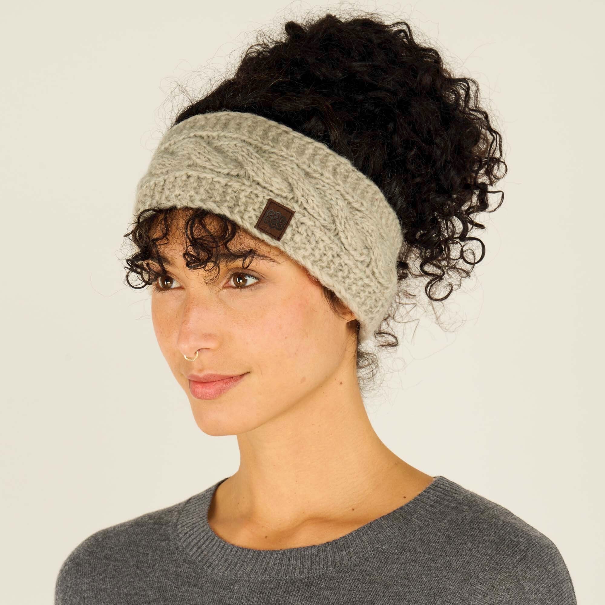 The same woman is shown in a three-quarter profile view, wearing the same thick, Sherpa Adventure Gear Kunchen Headband in Light Brown with a cable-knit pattern. Her curly dark hair is pulled up into a voluminous bun, and the headband covers her ears. The small, square, brown leather patch with a logo is visible on the side of the headband. She is wearing the same dark gray sweater. Her expression is calm and pleasant, with a slight smile, and she gazes to the side. The background remains neutral beige.