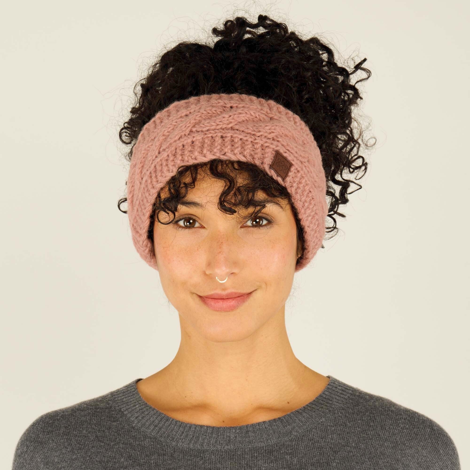 A woman with medium skin tone, dark curly hair tied up into a loose bun, and a small nose ring, is wearing a Sherpa Adventure Gear Kunchen Headband in Pink with a braided cable knit pattern. The headband covers her ears and the top of her forehead, leaving her curls visible above it. A small brown leather logo patch is sewn onto the left side of the headband. She is wearing a grey crewneck sweater and has a gentle smile while looking directly at the camera against a plain beige background.