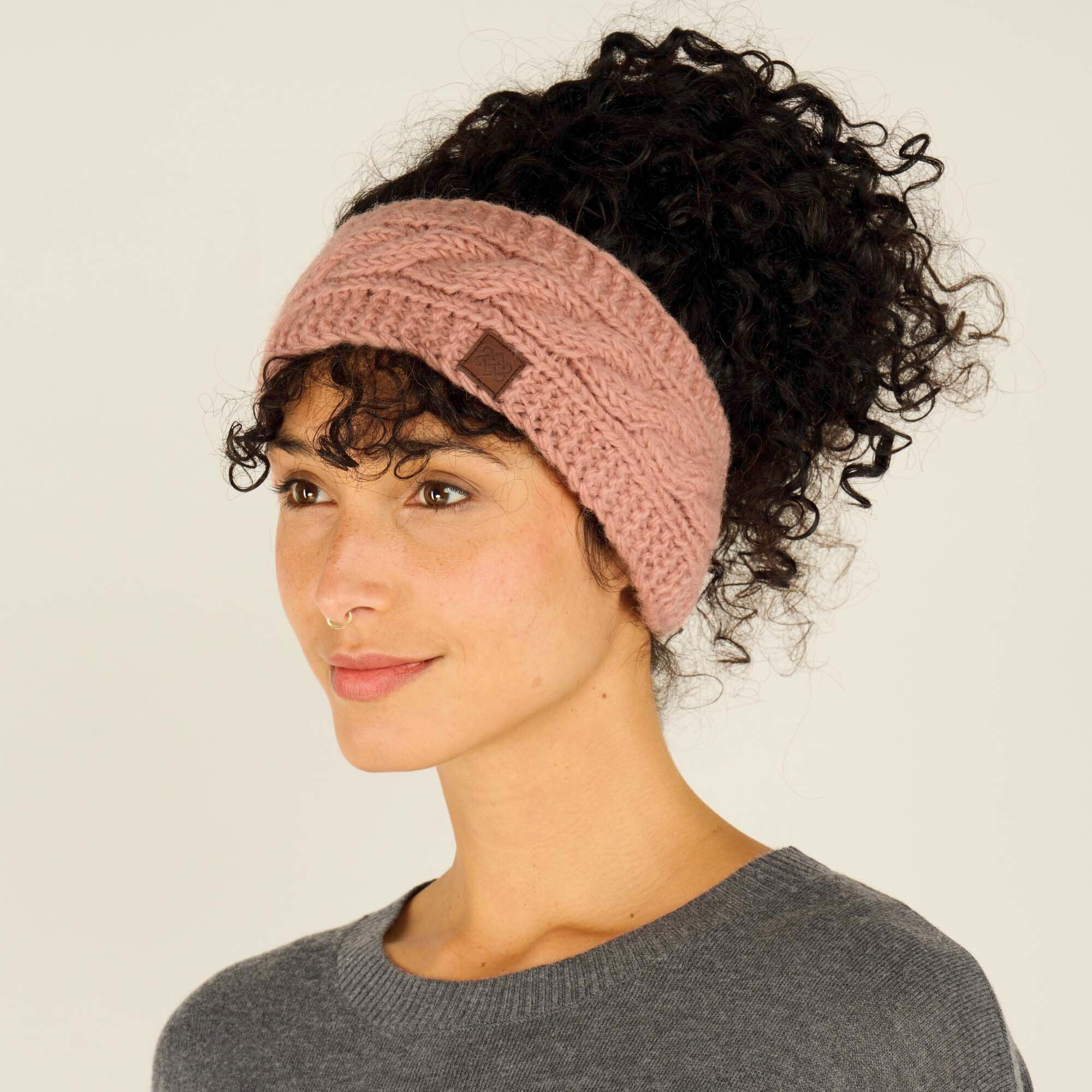 The same woman is shown from a three-quarter side angle, wearing the same Sherpa Adventure Gear Kunchen Headband in Pink. The braided cable knit pattern and the brown leather logo patch are clearly visible. Her dark curls are pulled up, showing how the headband fits around her head. She maintains a relaxed expression while looking off to the side. She is still wearing the grey crewneck sweater, and the background remains a plain beige.