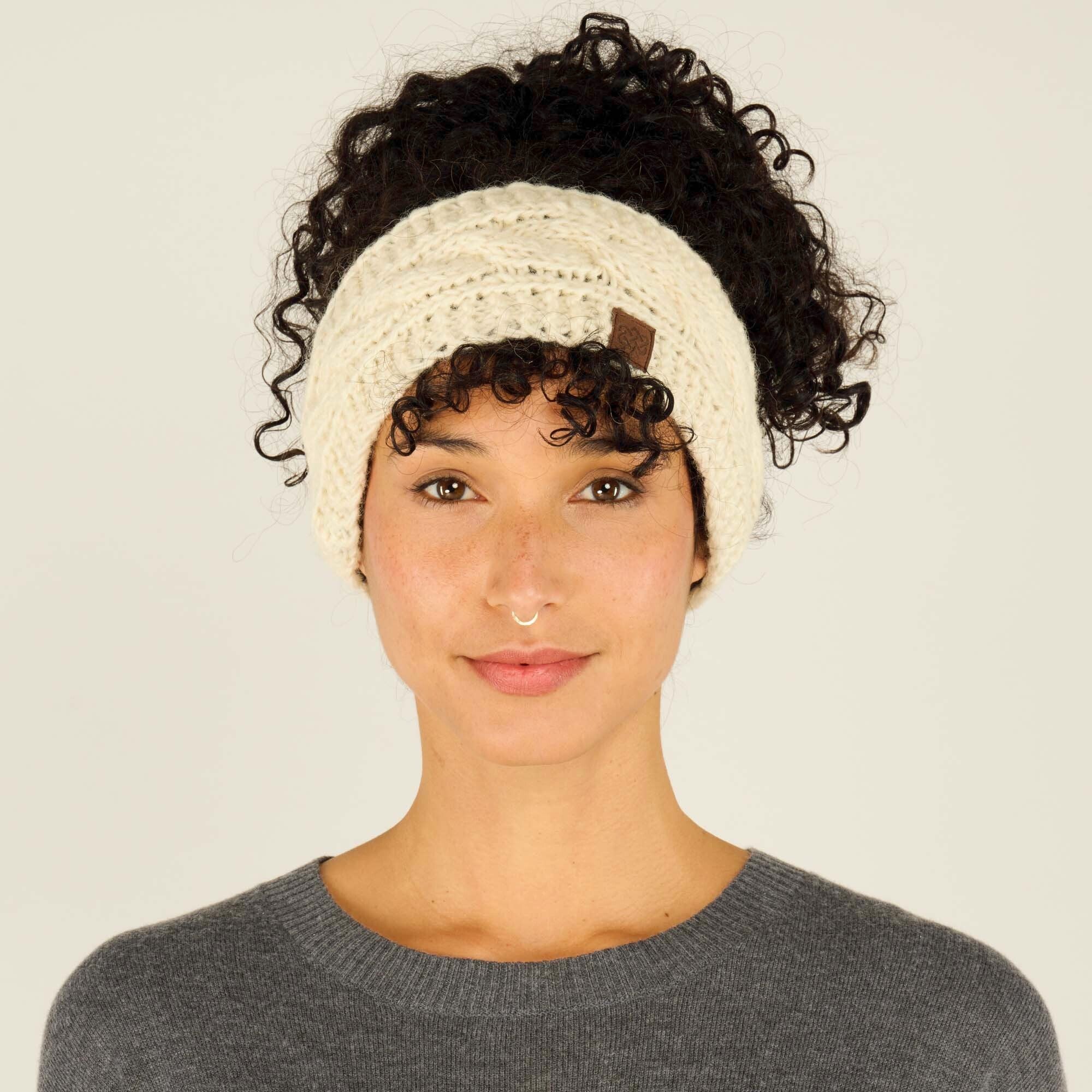 A woman with medium skin tone and voluminous curly black hair tied up in a high bun wears a Sherpa Adventure Gear Kunchen Headband in White. The headband features a braided cable knit design and a small rectangular brown logo patch on the left side of her forehead. She has a neutral expression with a soft smile and wears a gray sweater. The background is plain off-white.