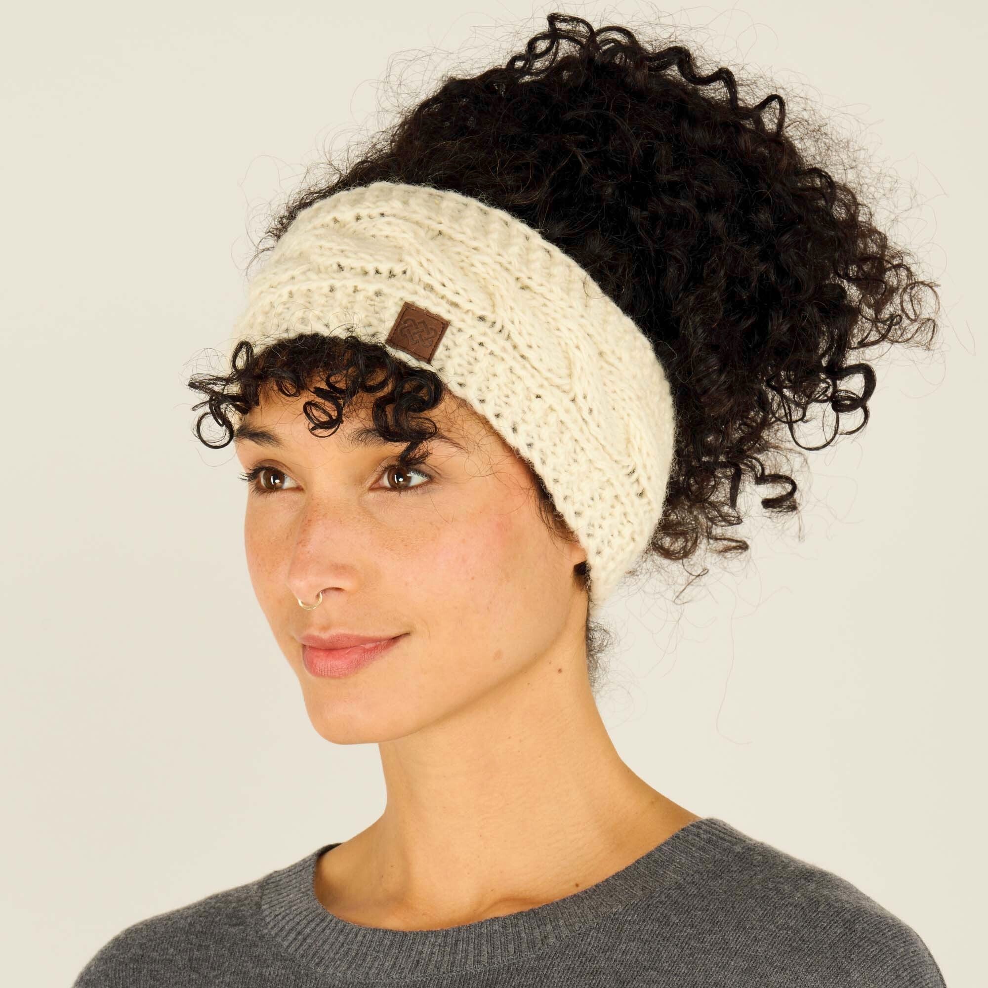 A side profile view of the same woman wearing the Sherpa Adventure Gear Kunchen Headband in White. The braided cable knit pattern and the small brown logo patch are visible. Her curly hair cascades down from the bun, and she looks off to the side with a relaxed expression. The gray sweater and plain off-white background are consistent with the first image.