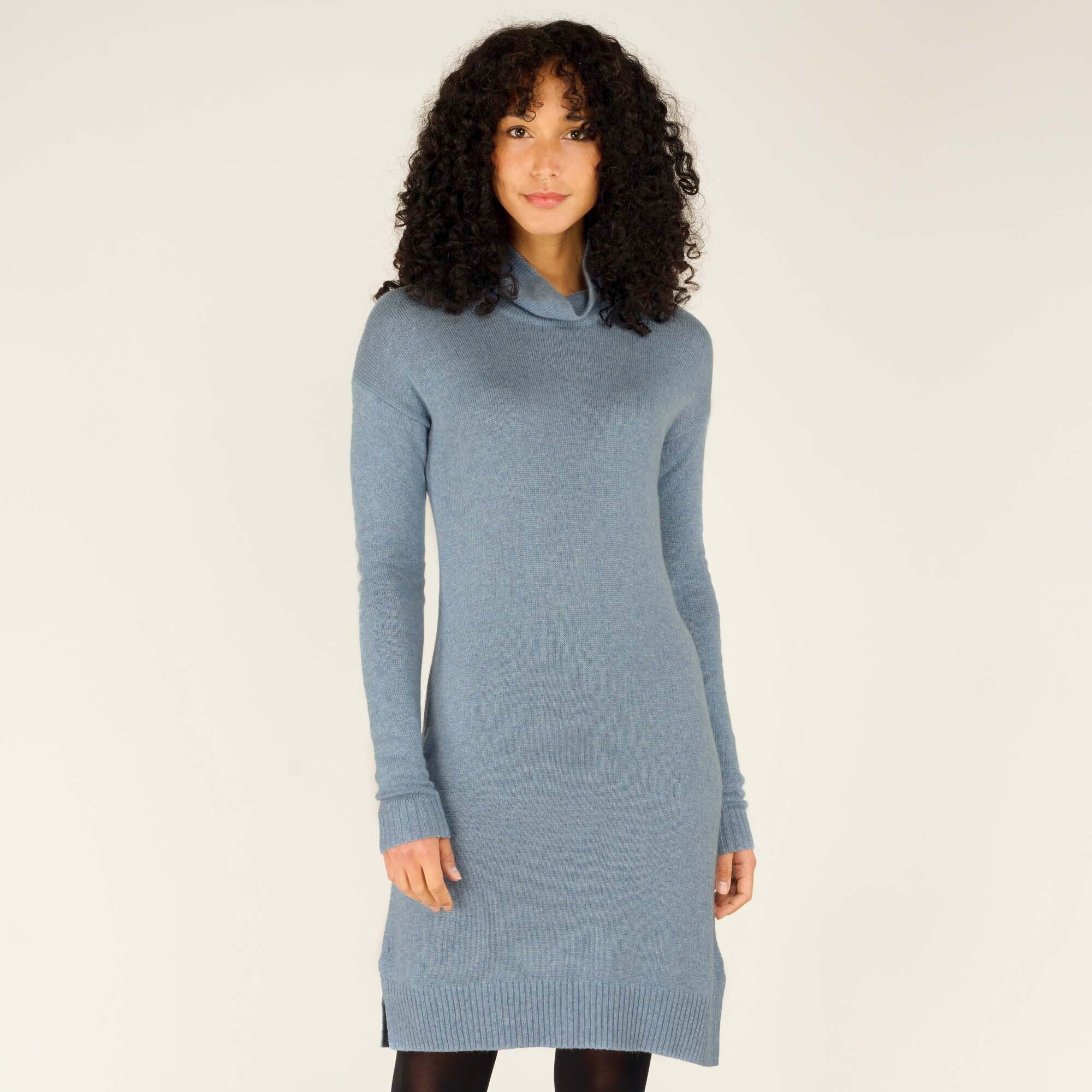 A model wearing a soft blue knit dress with a Sherpa Adventure Gear Lhakpa Mock Neck Dress in Blue and long sleeves, standing against a neutral background. The dress falls just above the knees, features ribbed cuffs, and has a subtle side slit near the hem. She pairs it with black tights and ankle boots for a minimalist and polished look.