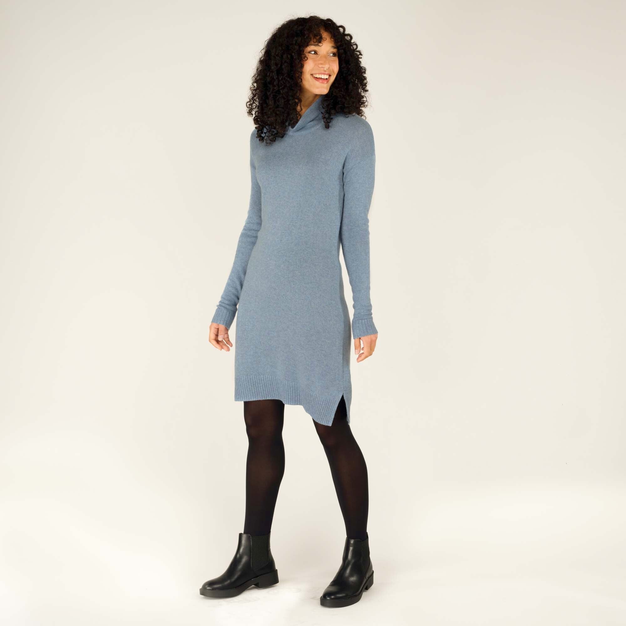 The model is captured in a three-quarter pose, showcasing the soft texture and figure-flattering fit of the Sherpa Adventure Gear Lhakpa Mock Neck Dress in Blue. Her curly hair frames her face as she smiles warmly. The side slit and ribbed hem detail are subtly visible.