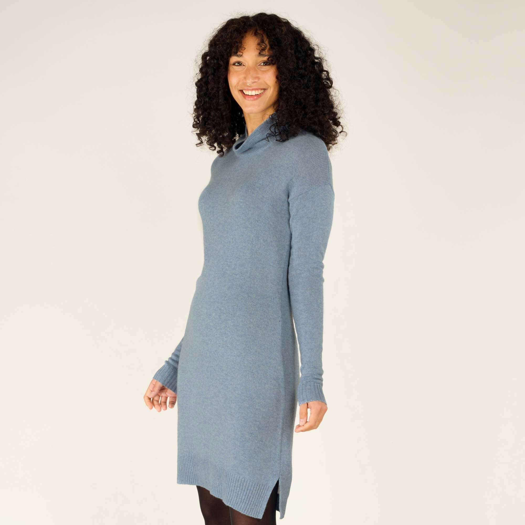 A closer side view of the model in the Sherpa Adventure Gear Lhakpa Mock Neck Dress in Blue, highlighting the smooth finish of the fabric and the drape of the dress along her silhouette. The long sleeves and ribbed edges are clearly detailed in this shot.