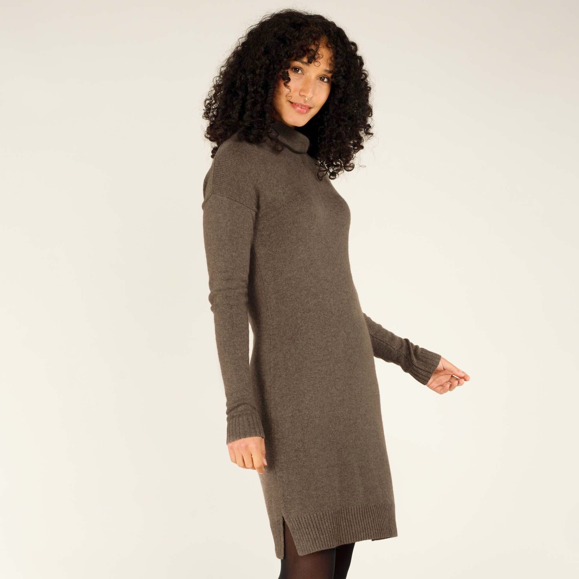 A side profile view of the woman wearing the Sherpa Adventure Gear Lhakpa Mock Neck Dress in Grey. The image captures the smooth contours of the fabric and the subtle side slits at the hemline. She glances back over her shoulder with a soft expression.