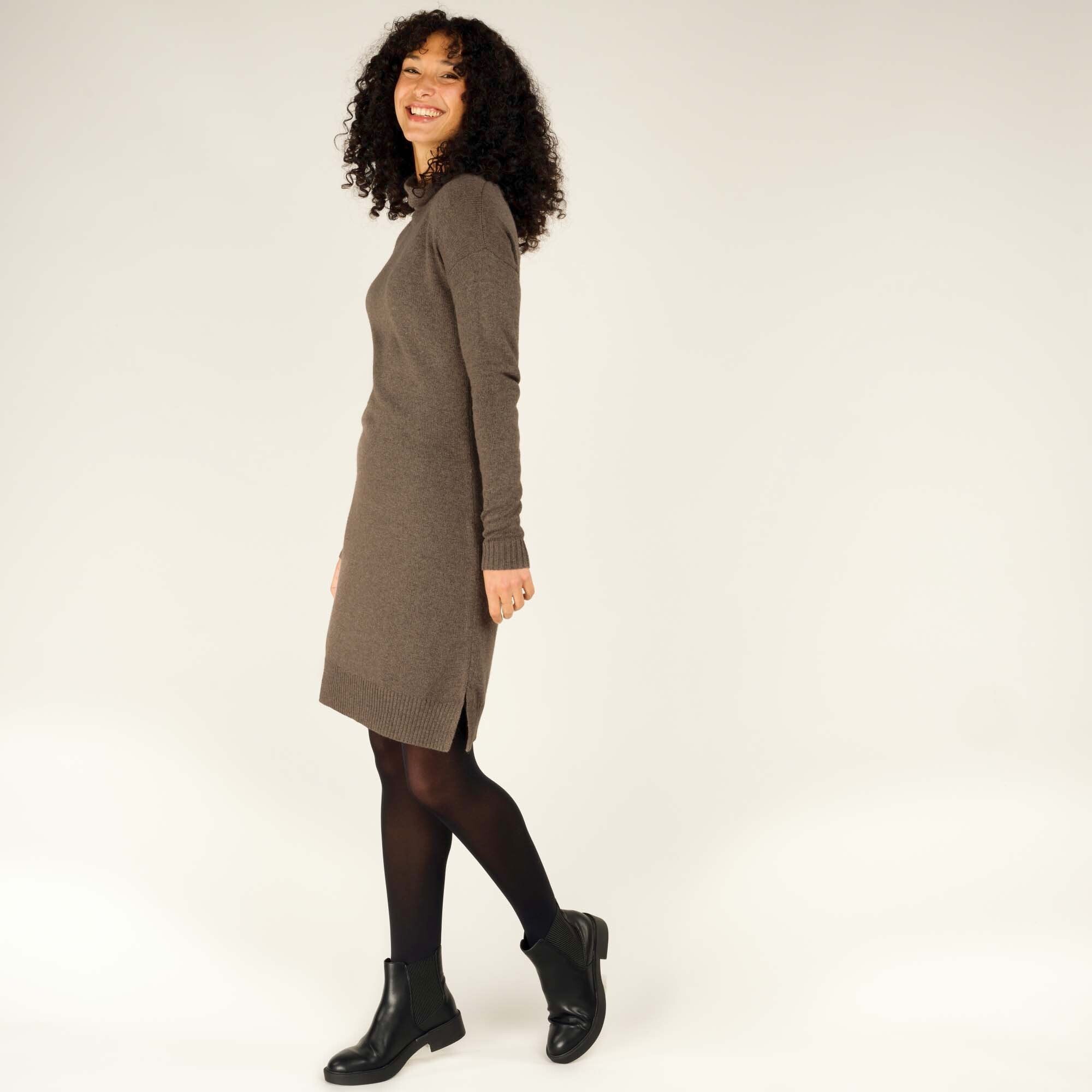 The model is mid-step, adding motion to the photo, as she smiles and looks toward the camera. The Sherpa Adventure Gear Lhakpa Mock Neck Dress in Grey and black tights are paired with sleek black ankle boots, demonstrating the outfit's versatility and comfort in movement.