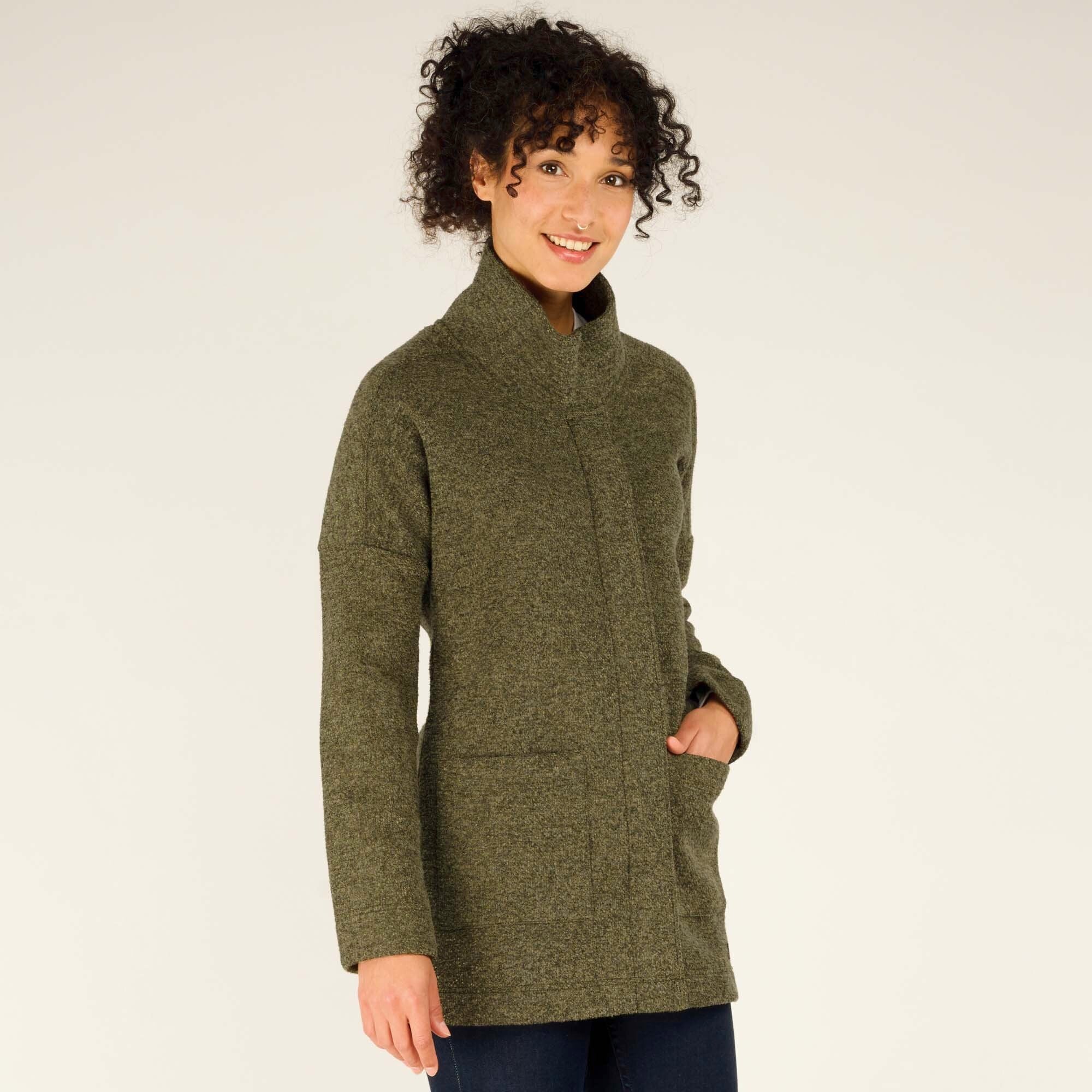 A woman is modeling a Sherpa Adventure Gear Lhamu Coat in Green with a high collar and concealed button closure. She is standing slightly turned to the side, her hands in the coat pockets, smiling softly. The coat has a subtle textured fabric, two front pockets, and a tailored fit.