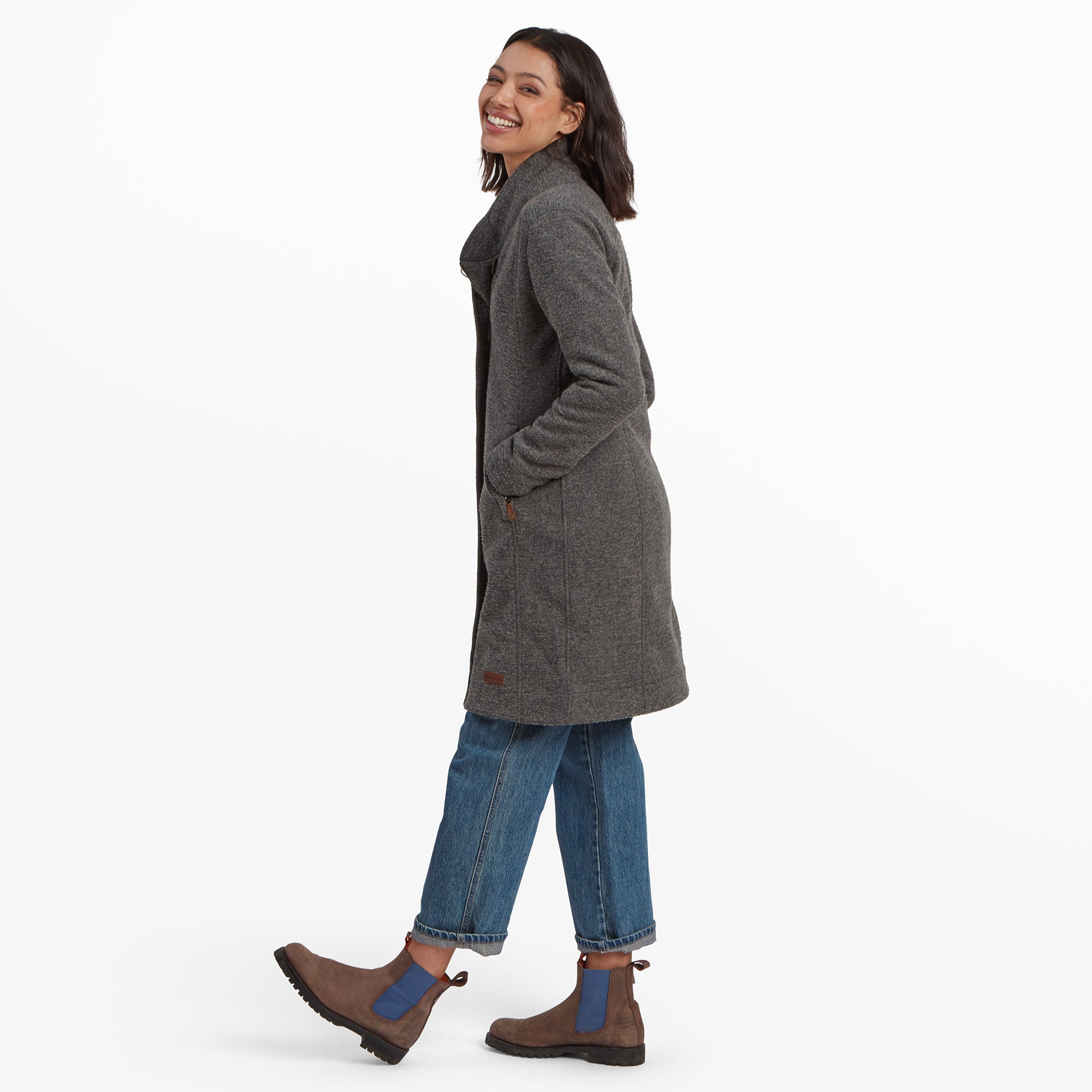 The same woman poses with her hands gently touching the collar of her Sherpa Adventure Gear Lhamu Jacket in Grey. The coat has an elegant, structured fit with discreet side pockets featuring brown leather pull tabs. The outfit is composed of a beige sweater, blue jeans, and brown ankle boots.