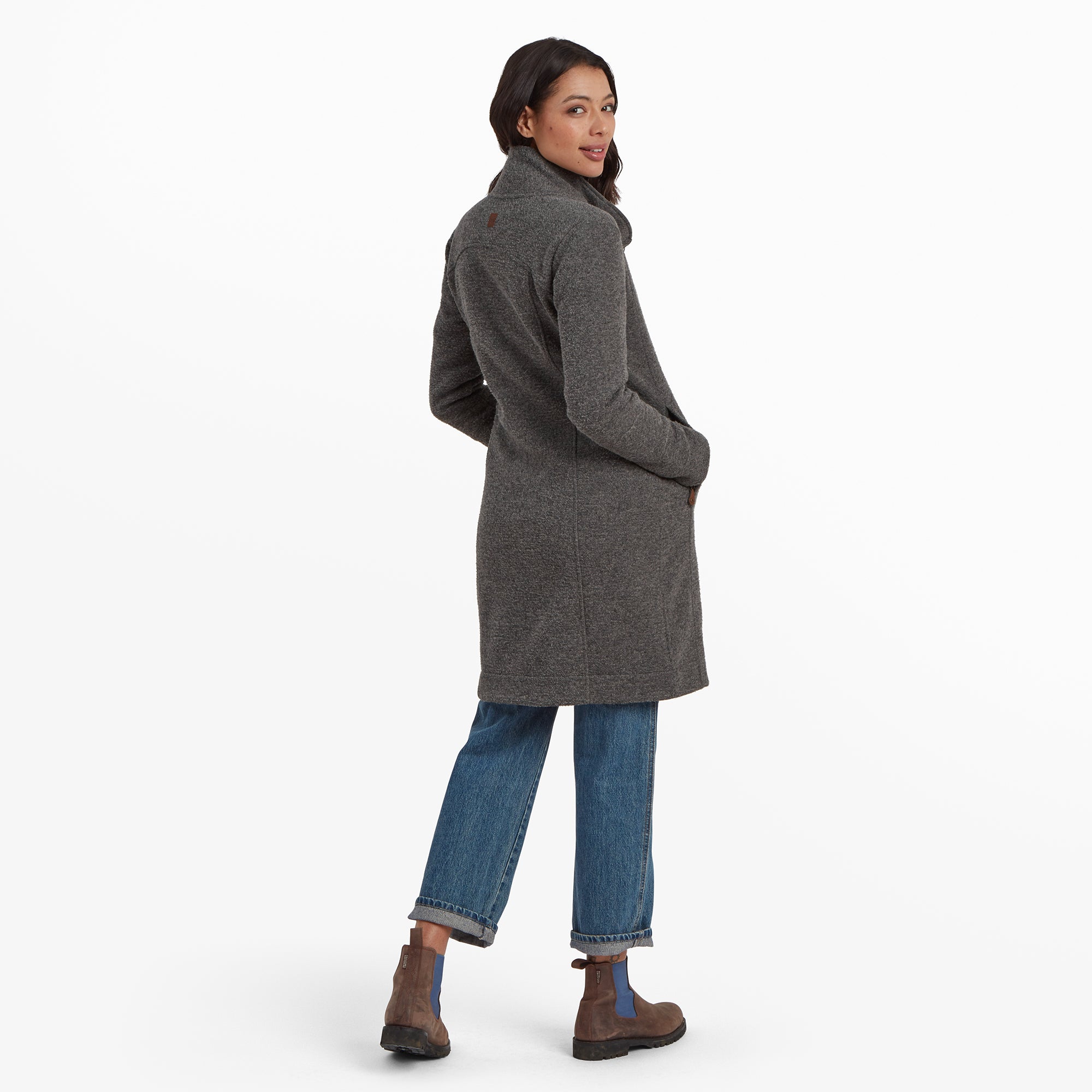 She is captured in a candid moment, smiling while looking sideways. The Sherpa Adventure Gear Lhamu Jacket in Grey comfortably, showing its mid-thigh length and a slightly curved hem. Her jeans are cuffed at the ankles, revealing the full height of her brown boots with blue panels.