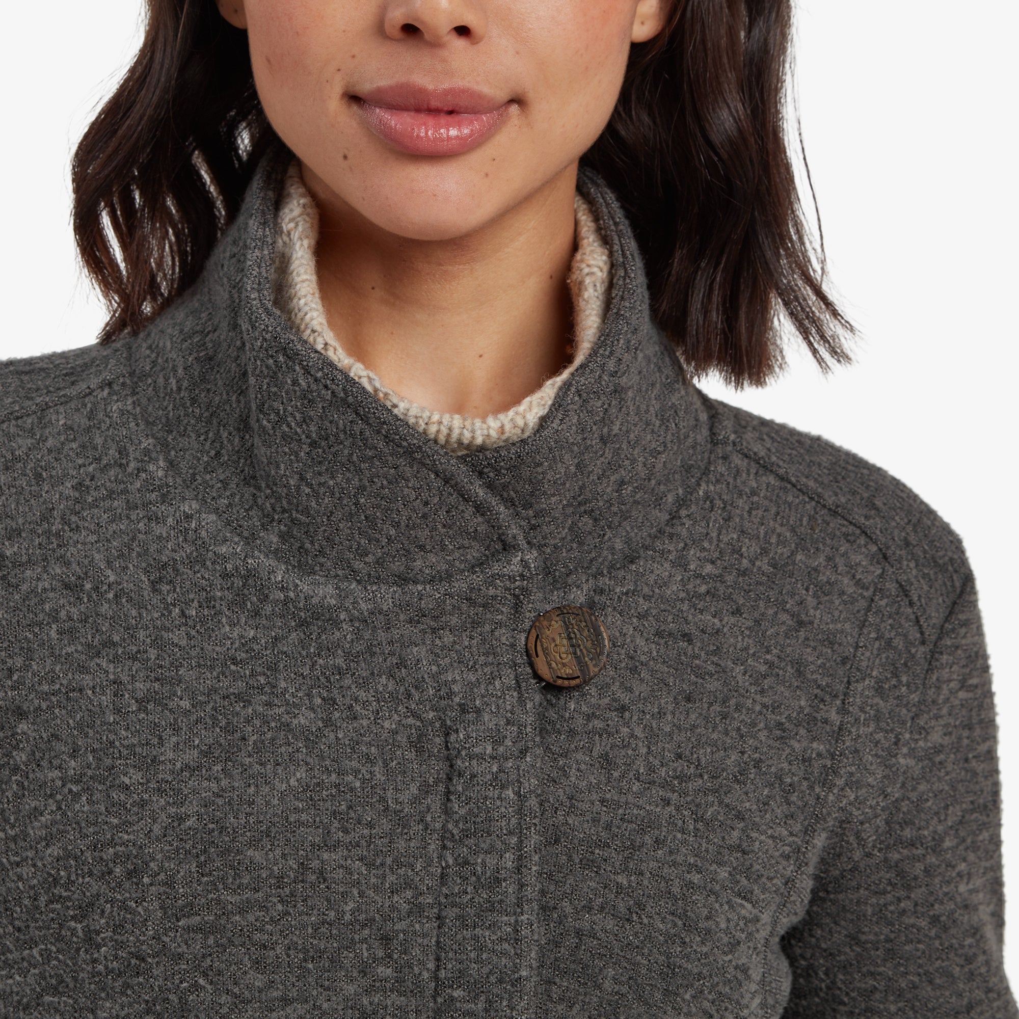 The back of the Sherpa Adventure Gear Lhamu Jacket in Grey is showcased, revealing its seamless design and structured collar. The woman's pose with her hands tucked into the pockets adds a relaxed feel. She pairs the coat with blue jeans and brown boots.