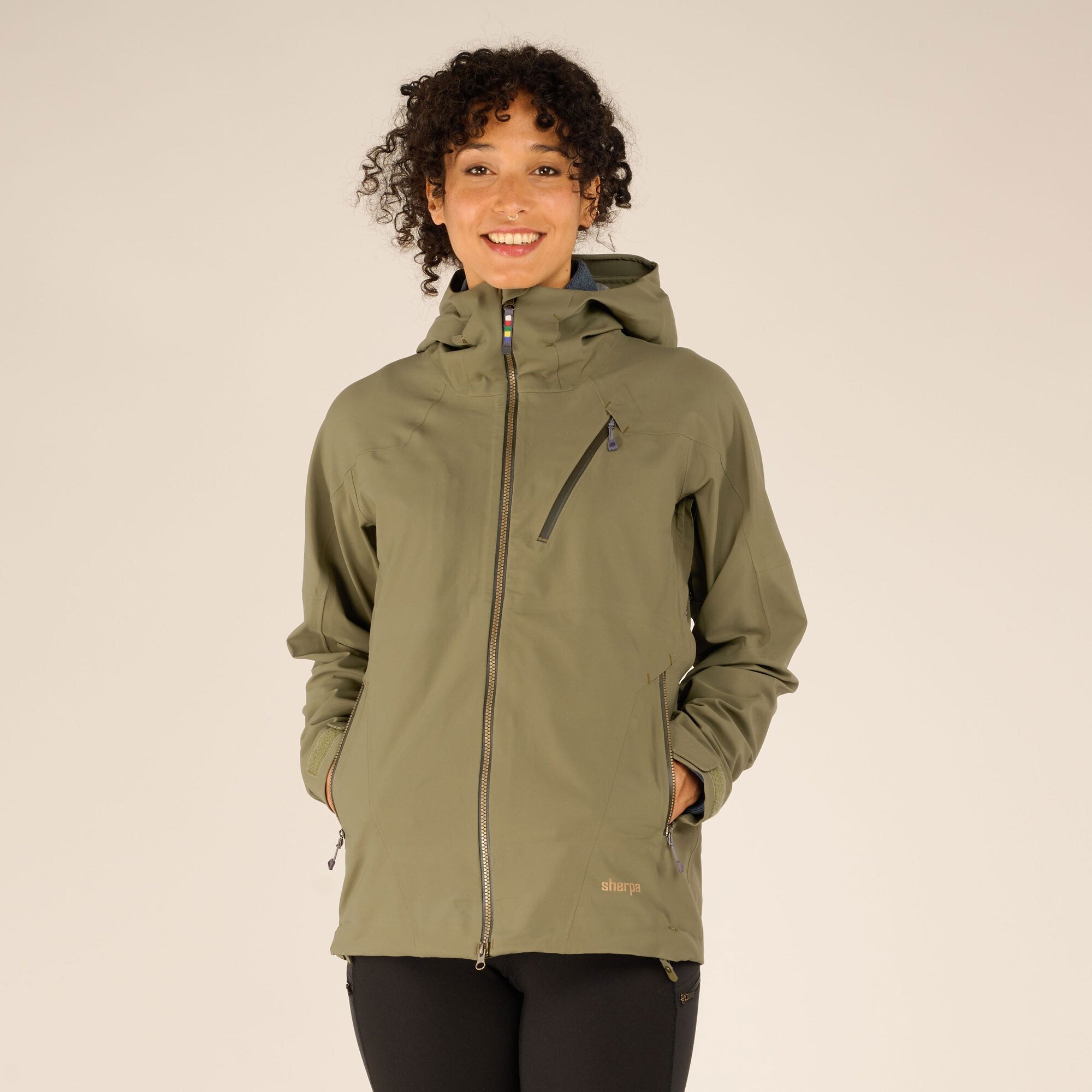 A smiling model wearing a Sherpa Adventure Gear Makalu Eco Jacket in Green, standing with her hands in the side pockets. The jacket features a zippered chest pocket, a high collar, and a fitted hood. The background is light beige.