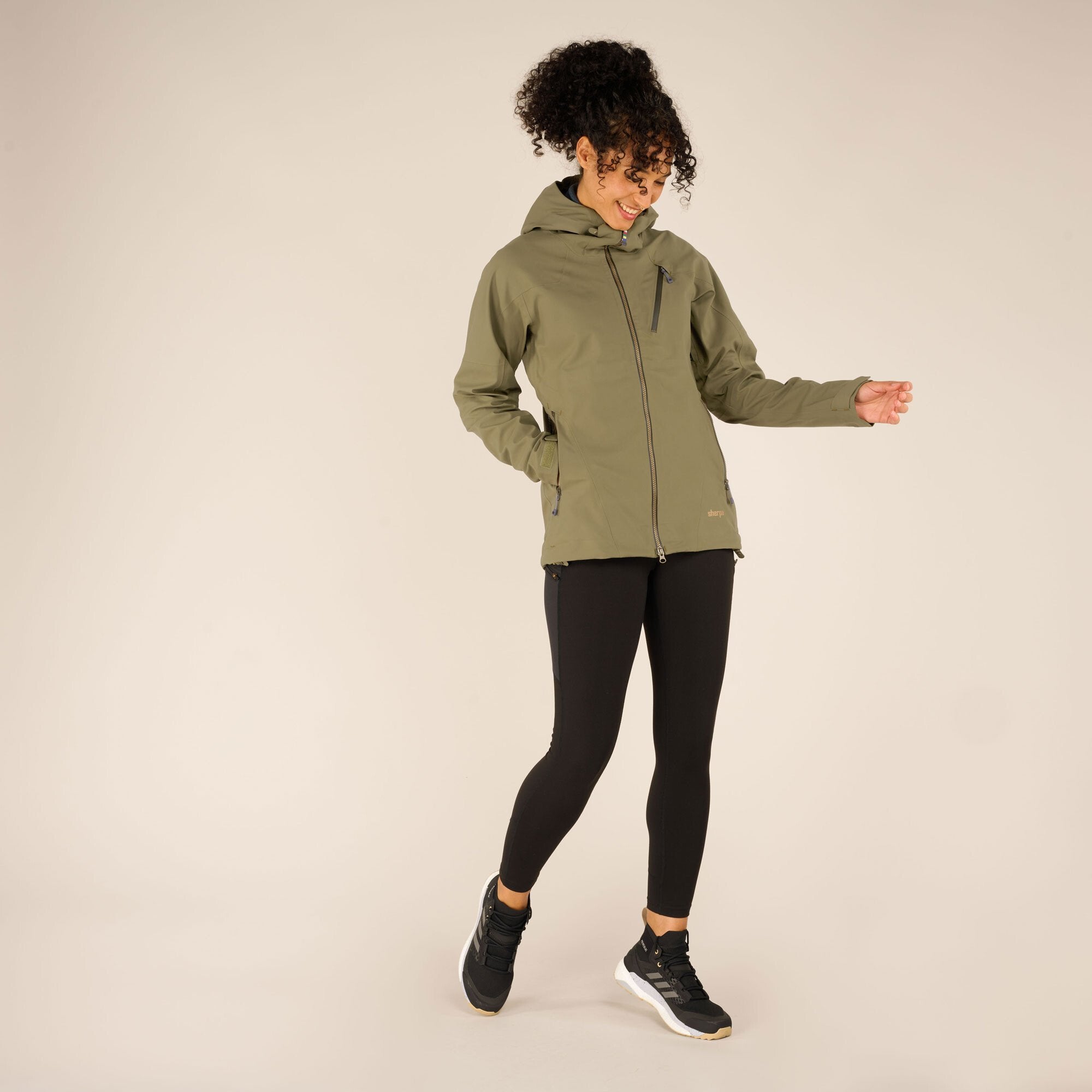 Full-body view of the model in the Sherpa Adventure Gear Makalu Eco Jacket in Green paired with black leggings and black trainers. The model stands in a slightly playful pose, holding the jacket's hem with one hand while smiling.