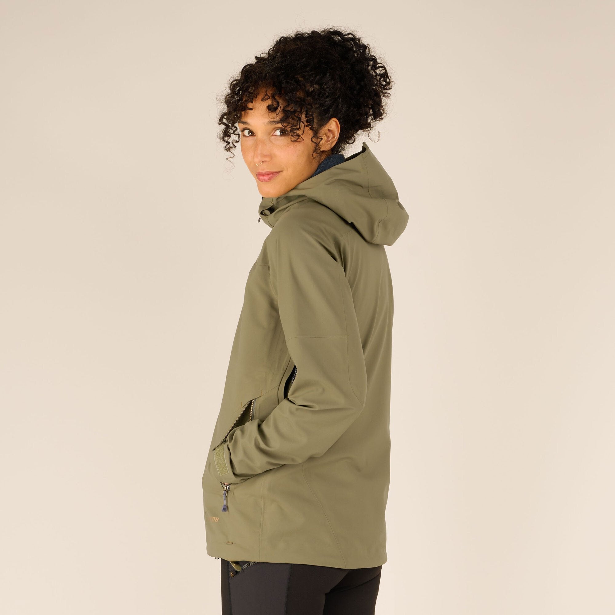 A side view of the model in the Sherpa Adventure Gear Makalu Eco Jacket in Green, showcasing the contoured fit and the hood lying flat on the back. Her hand is casually placed in the jacket’s side pocket, highlighting its functional design.