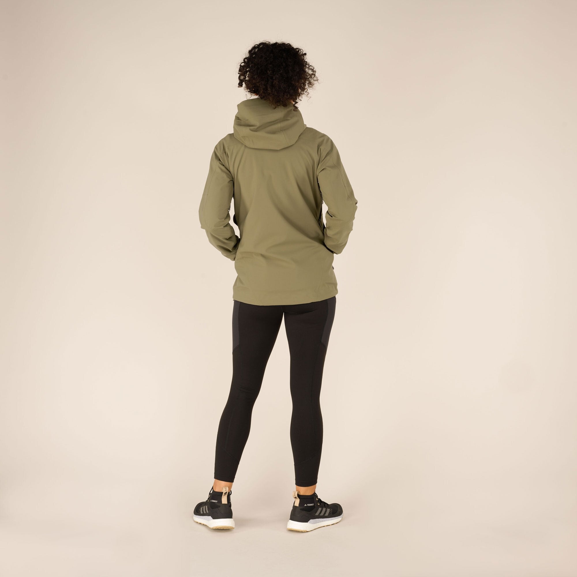 Rear view of the model wearing the Sherpa Adventure Gear Makalu Eco Jacket in Green, emphasising the clean back design and the extended hem for extra coverage. The hood is neatly resting on the model's back. She is wearing black leggings and trainers.