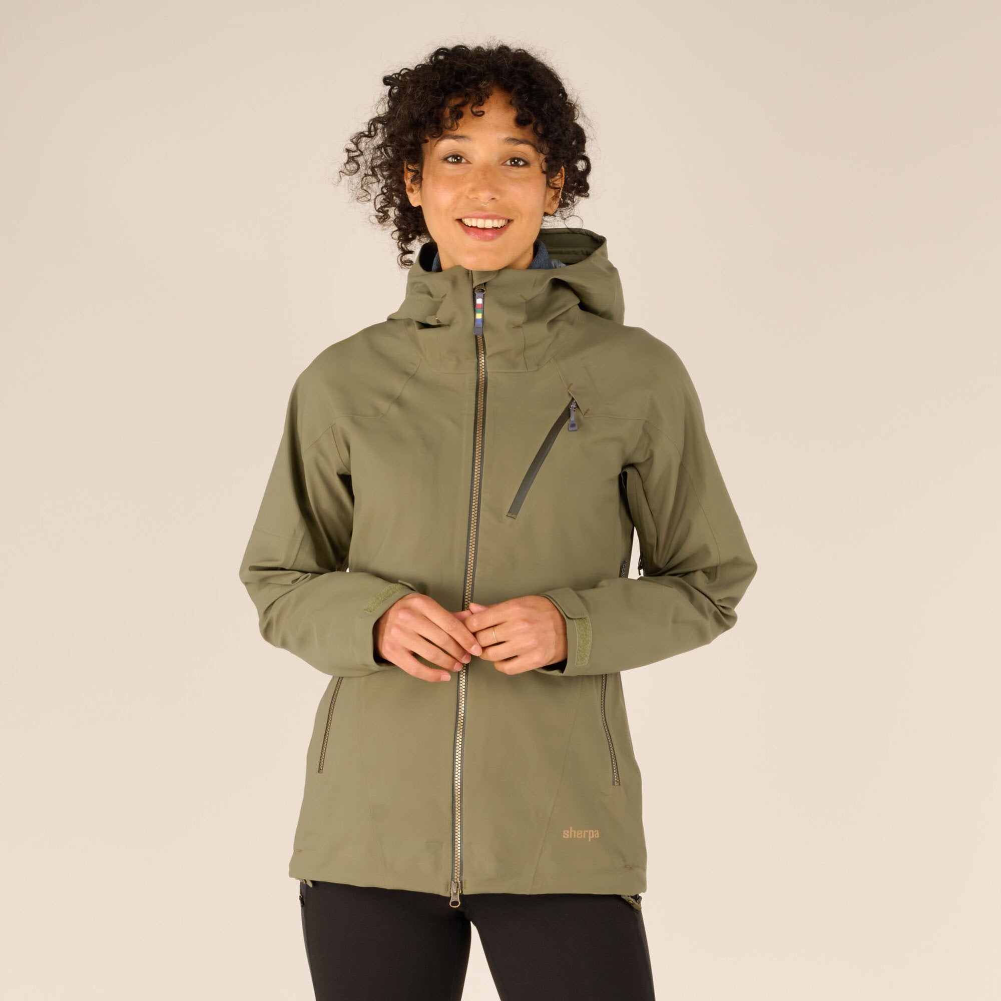 A close-up of the front of the Sherpa Adventure Gear Makalu Eco Jacket in Green, showing the model smiling while zipping the jacket fully to display its structured hood and front design. The Sherpa logo is subtly visible on the chest.