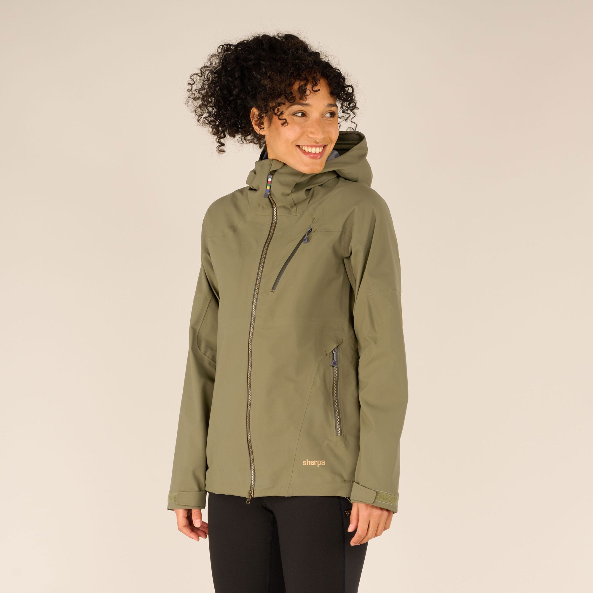 A three-quarter view of the model in the Sherpa Adventure Gear Makalu Eco Jacket in Green, smiling softly with her head tilted slightly. The jacket’s fitted design and functional zippers on the chest and side pockets are highlighted.