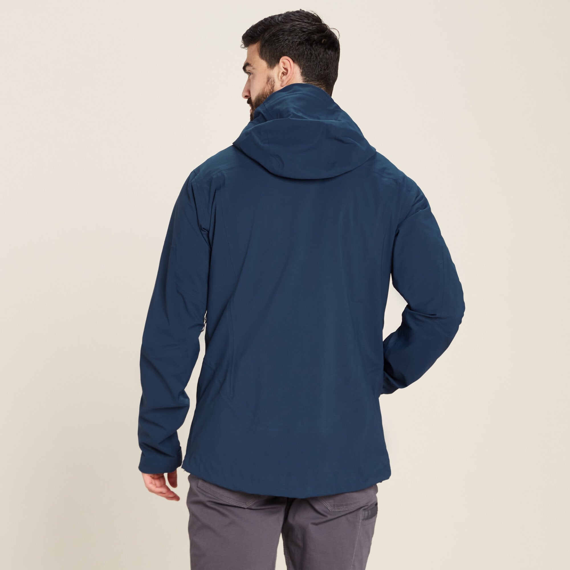 A rear view of a man wearing the Sherpa Adventure Gear Makalu Jacket in Blue. The hood is visible, lying flat on his back, showcasing the jacket's streamlined design. The model's arms are relaxed by his sides as he looks slightly to his left. The background is plain beige.