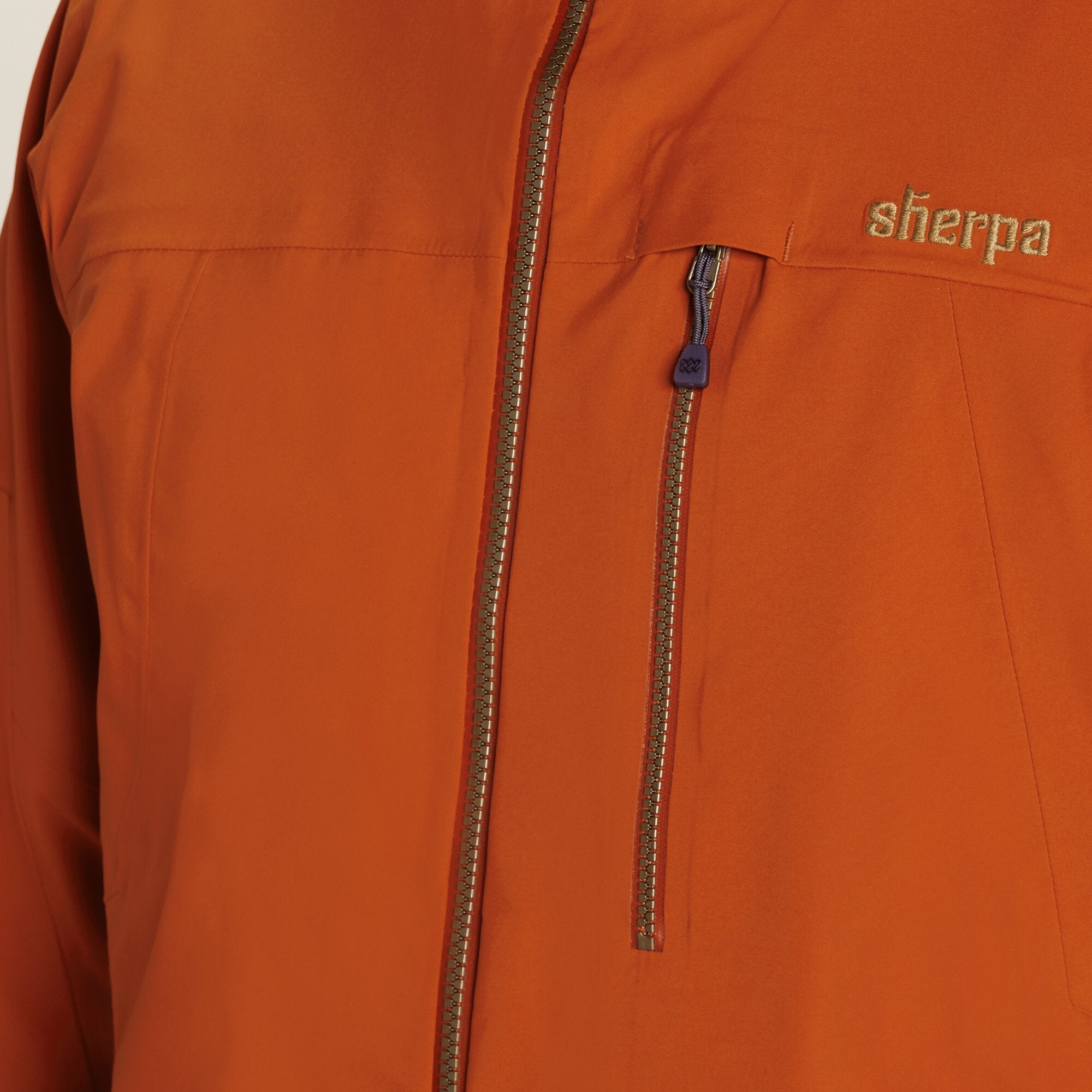 A detailed view of the chest area of a Sherpa Adventure Gear Makalu Jacket in Orange. The image highlights a zippered chest pocket with a dark pull tab, accented by fine stitching. Above the pocket, the "Sherpa" logo is embroidered in a gold-tone thread, showcasing brand detail.