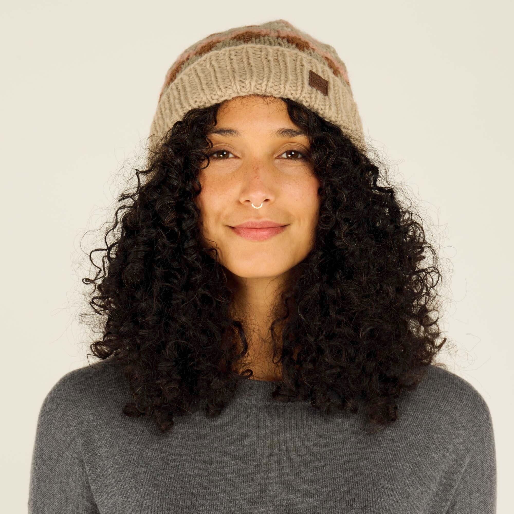 A close-up frontal view of a model wearing a Sherpa Adventure Gear Manaslu Hat in Beige with intricate zigzag patterns in brown, pink, and off-white hues. The model's natural curls frame their face, complementing the soft and cozy design of the hat. A small brown leather logo patch is visible on the front of the hat, adding a subtle branding detail.