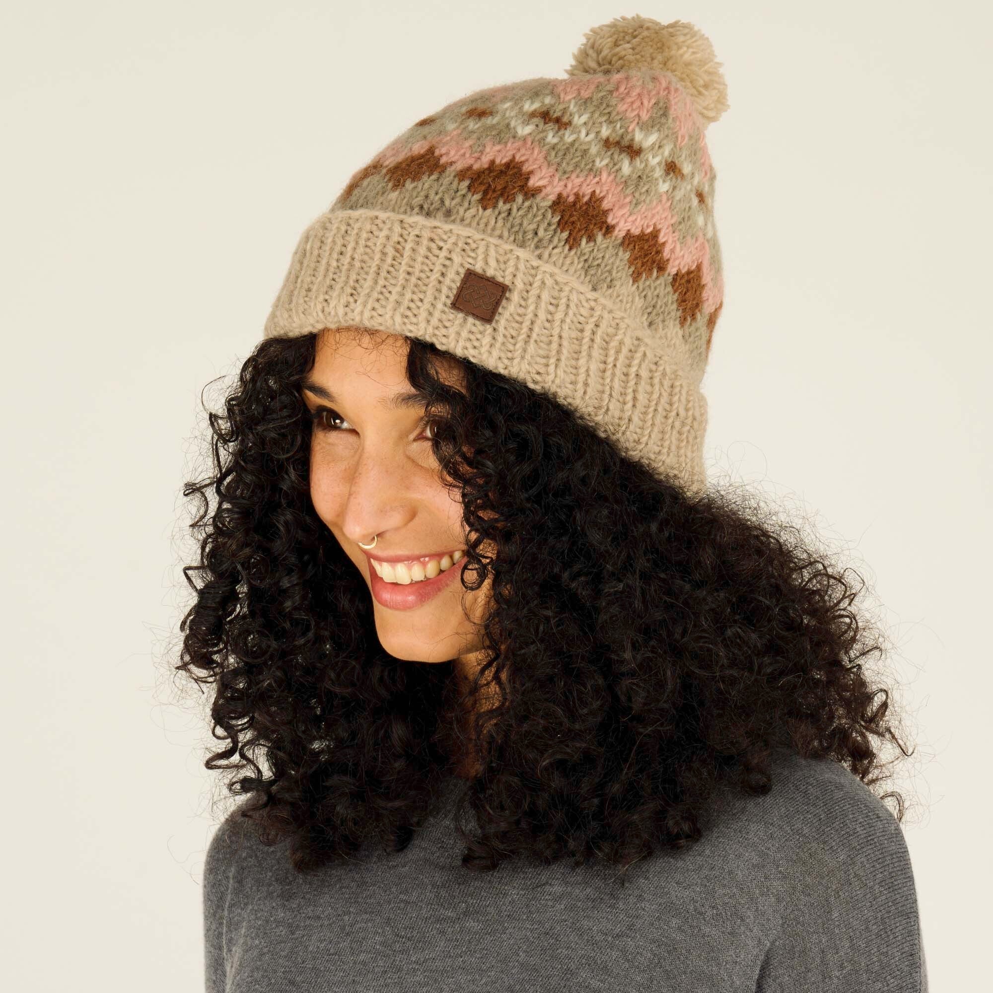 A side profile of the model smiling brightly while wearing the same Sherpa Adventure Gear Manaslu Hat in Beige with zigzag patterns and a playful pom-pom at the top. The texture of the hat and its snug fit are highlighted, with the pom-pom adding a whimsical touch to the overall design. The model's expression exudes warmth and comfort.