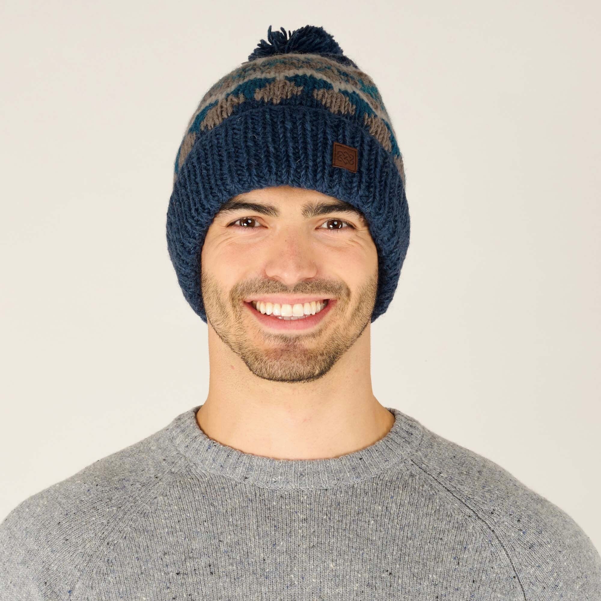 A smiling individual is wearing a cozy, Sherpa Adventure Gear Manaslu Hat in Blue adorned with a decorative pom-pom on top. The beanie features a textured knit pattern with muted blue, grey, and beige tones in a geometric design. A small square leather patch with subtle branding is visible on the front of the hat, adding a touch of style. The individual is also dressed in a light grey sweater with a speckled texture, complementing the hat’s casual and winter-ready aesthetic.