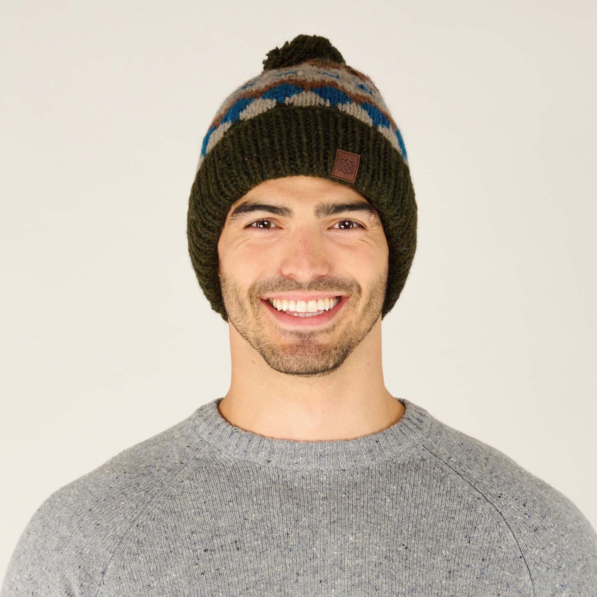 A front-facing close-up of the same male model, wearing the same Sherpa Adventure Gear Manaslu Hat in Green with a blue, beige, and brown geometric design. The brown leather patch is clearly visible on the brim. The model is smiling warmly, showcasing the beanie's snug fit and cozy texture. He is wearing a light gray knit sweater, and the background remains off-white and neutral for focus on the product.